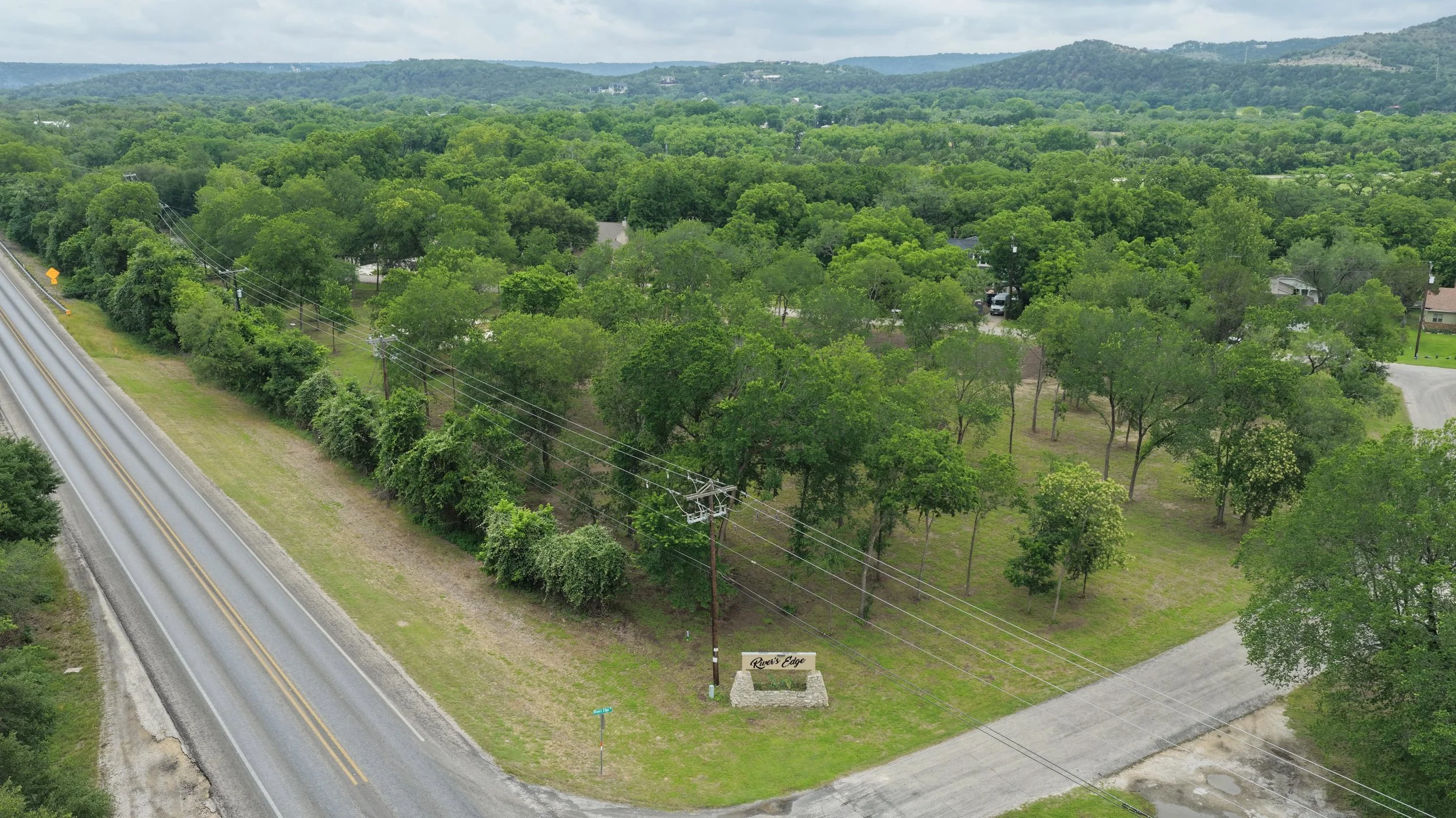 Aerial view of a landscape with a road, trees, and a sign that reads 'Rivers Edge' amid a green area, with hills in the background and cloudy sky.