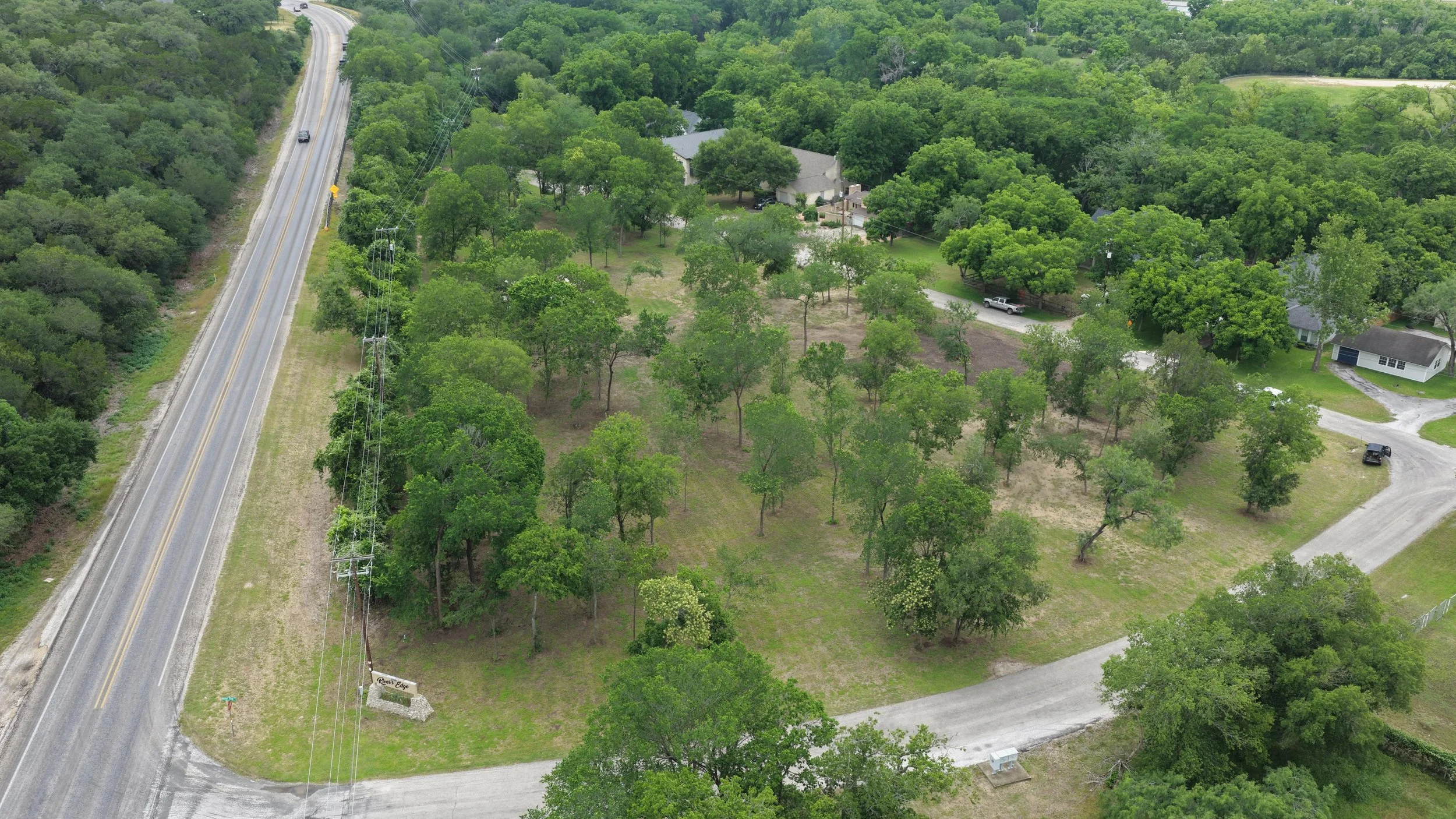 An aerial view of a residential area surrounded by lush green trees, with a road running along the left side and a few cars parked near houses on the right. There is a sign that reads 'Renee's Edge' near the bottom left.