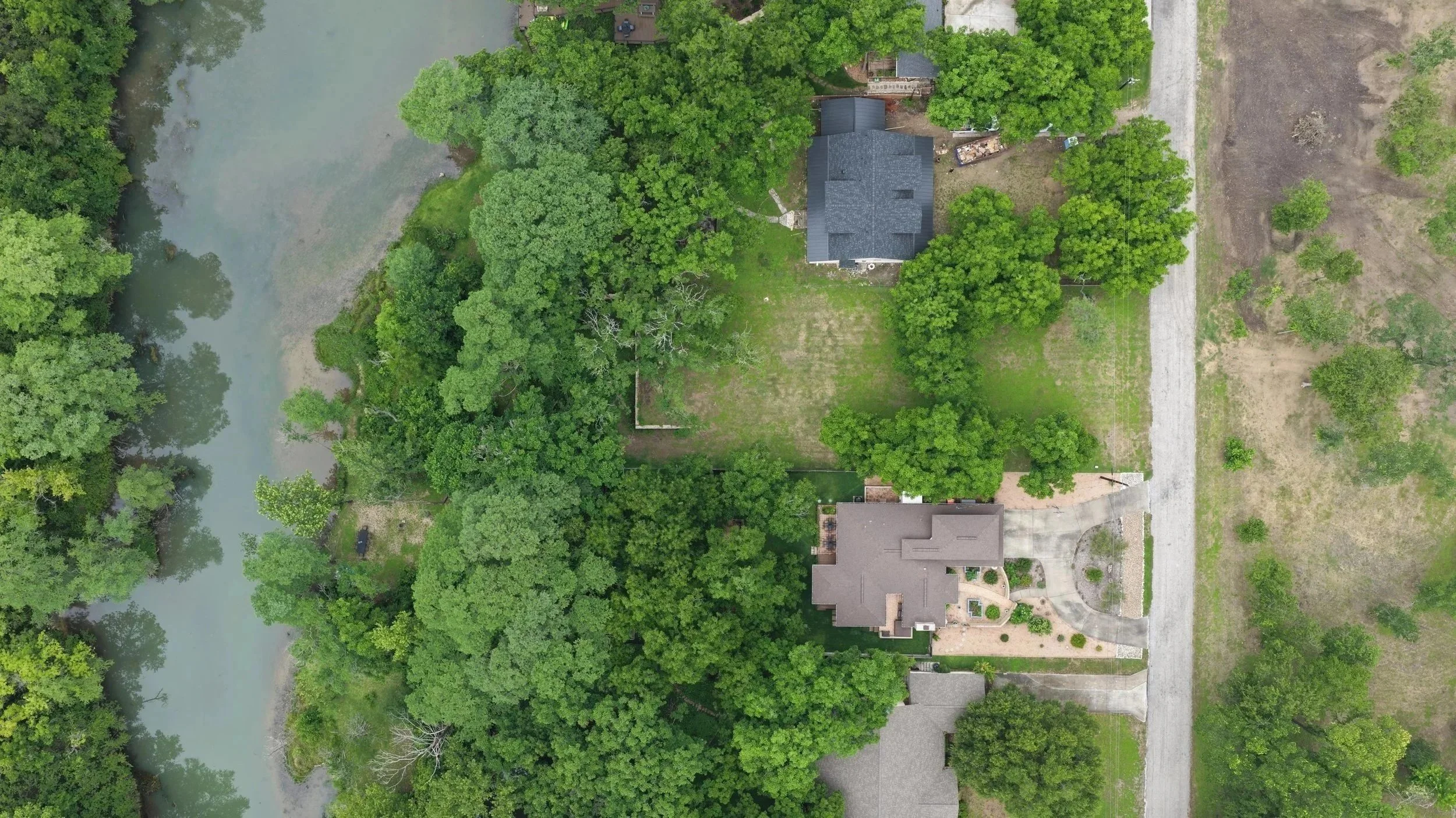 An aerial view of a house surrounded by dense green trees, with a partially visible river on the left side and a gravel road on the right side.