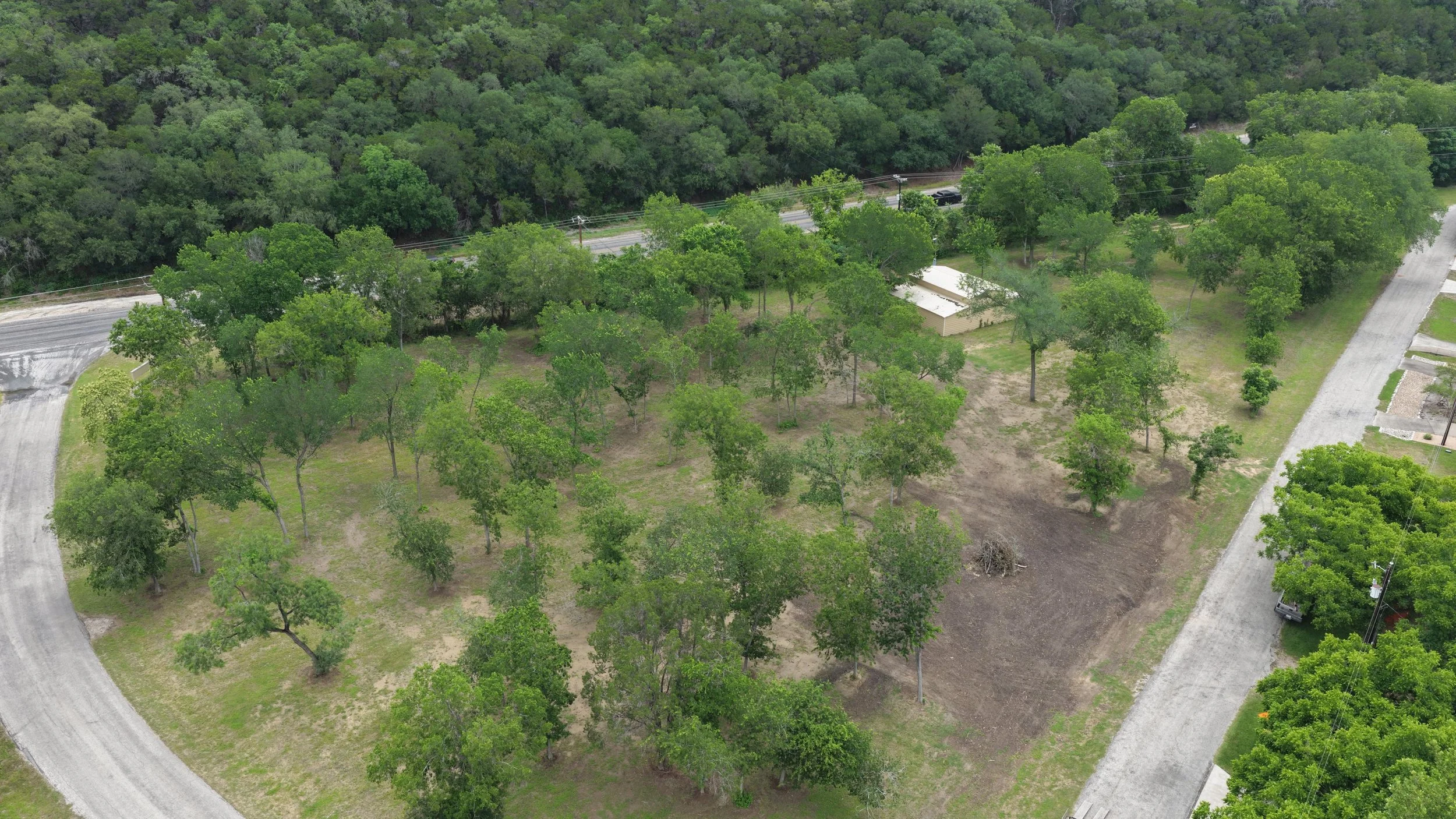 An aerial view of a rural scene with green trees, a small building, dirt and paved roads, and a densely wooded background.