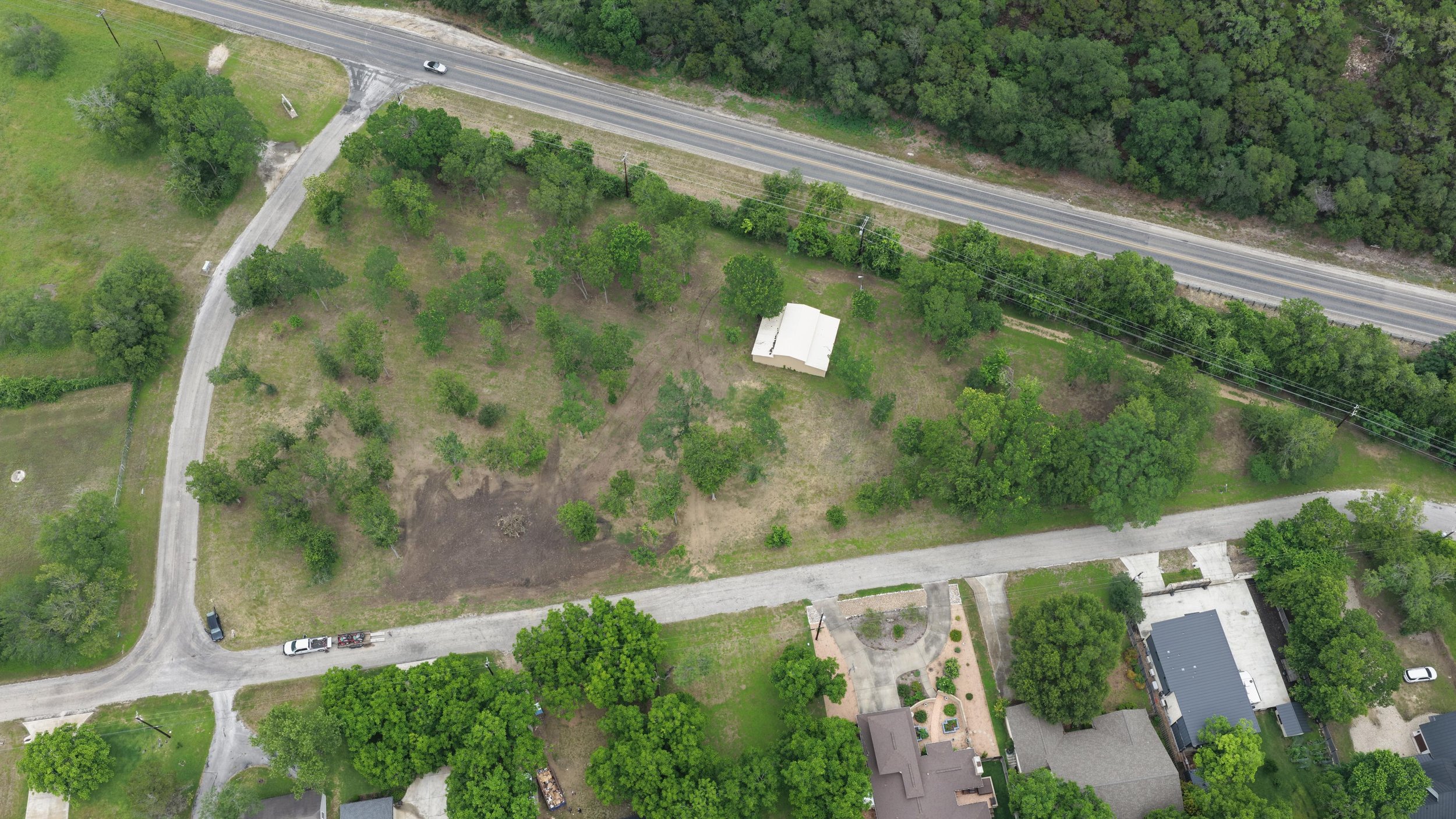 An aerial view of a residential area with a lot of greenery, including trees and lawns, a paved road, and a small building in the center surrounded by trees.