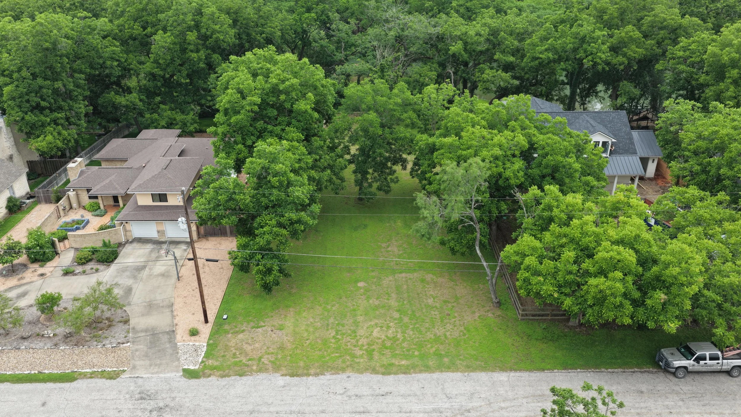 Aerial view of a suburban backyard with large trees, neighboring houses with patios, a driveway, and a grassy open space.