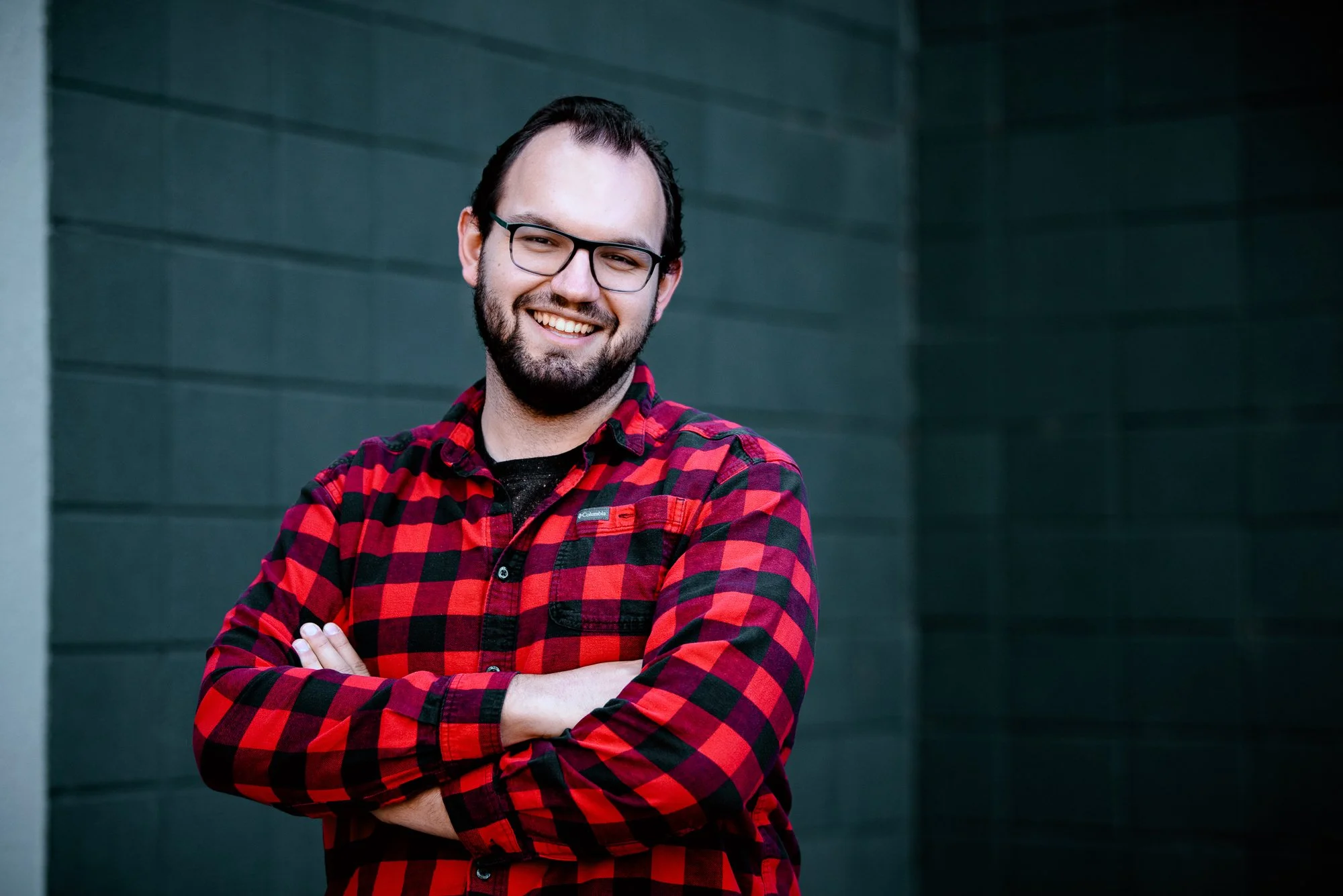 A man with glasses and a beard, smiling, wearing a red and black plaid shirt, standing with arms crossed in front of a dark green wall.