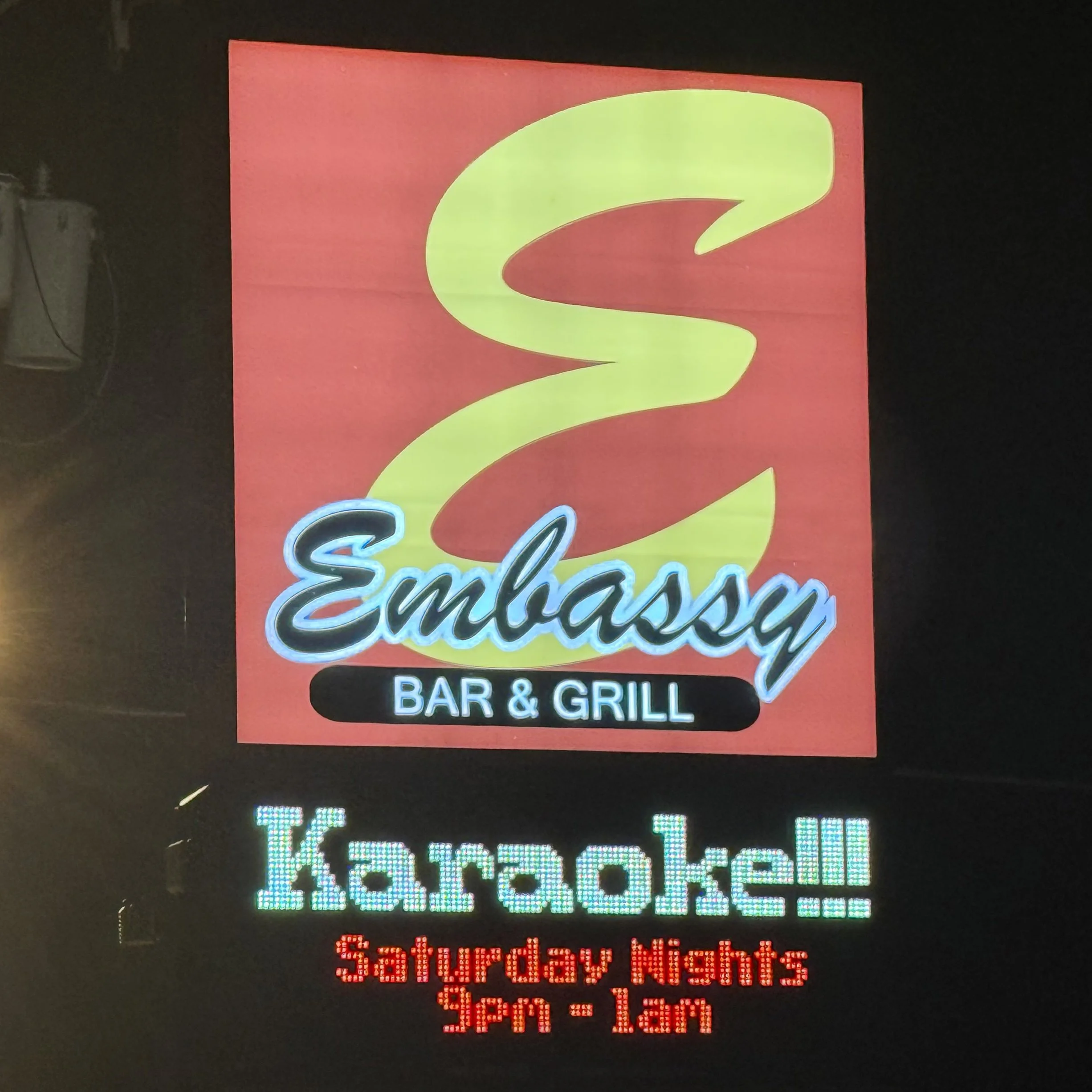 Illuminated Embassy Bar & Grill sign in West Olive, Michigan, advertising Saturday night karaoke from 9 PM to 1 AM during the GRkaraoke.com Karaoke Marathon 2026