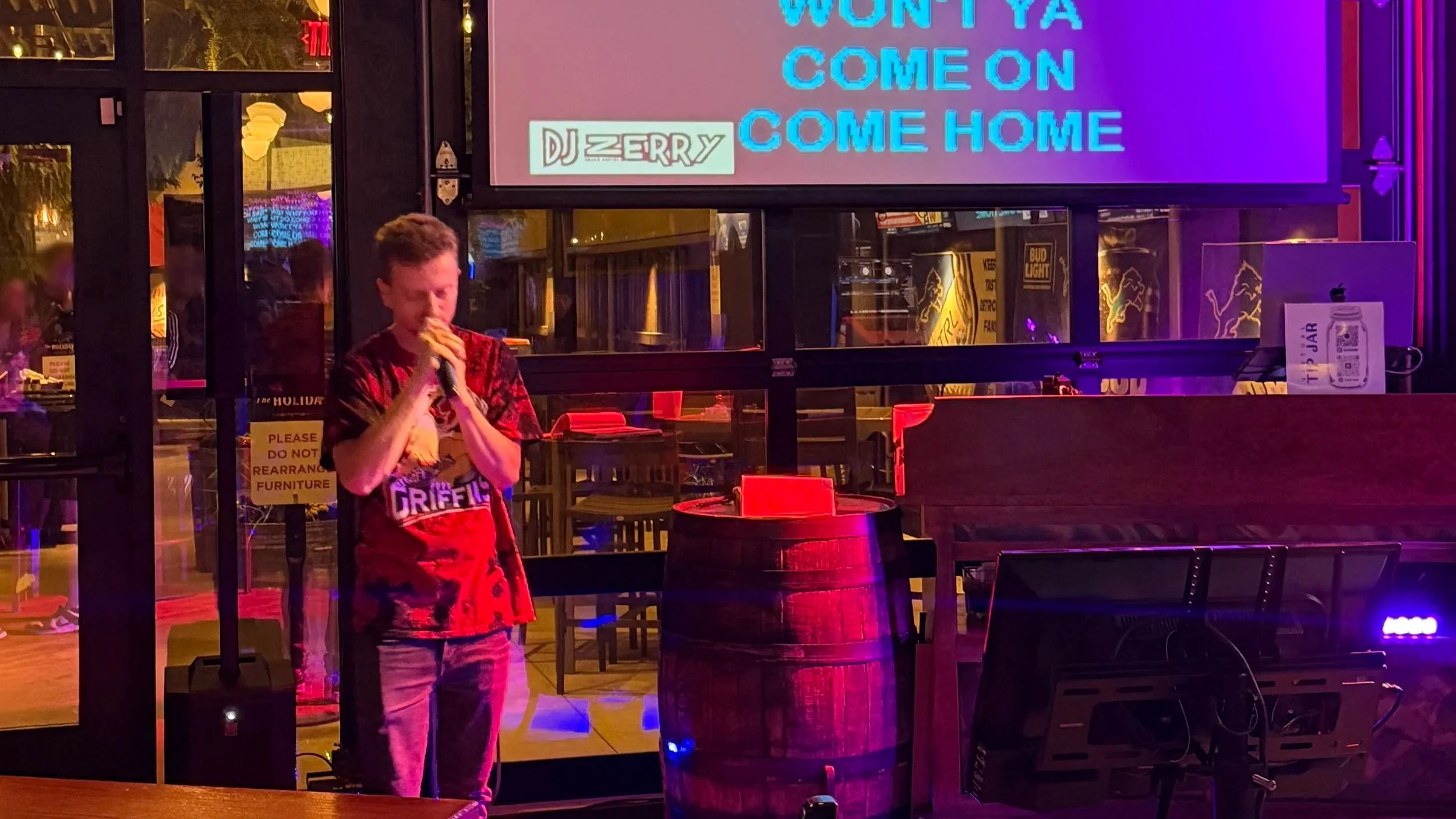 GRkaraoke.com founder Chris singing karaoke at The Holiday Bar on the West Side of Grand Rapids, Michigan, with projected lyrics and colorful stage lighting during the Karaoke Marathon 2026