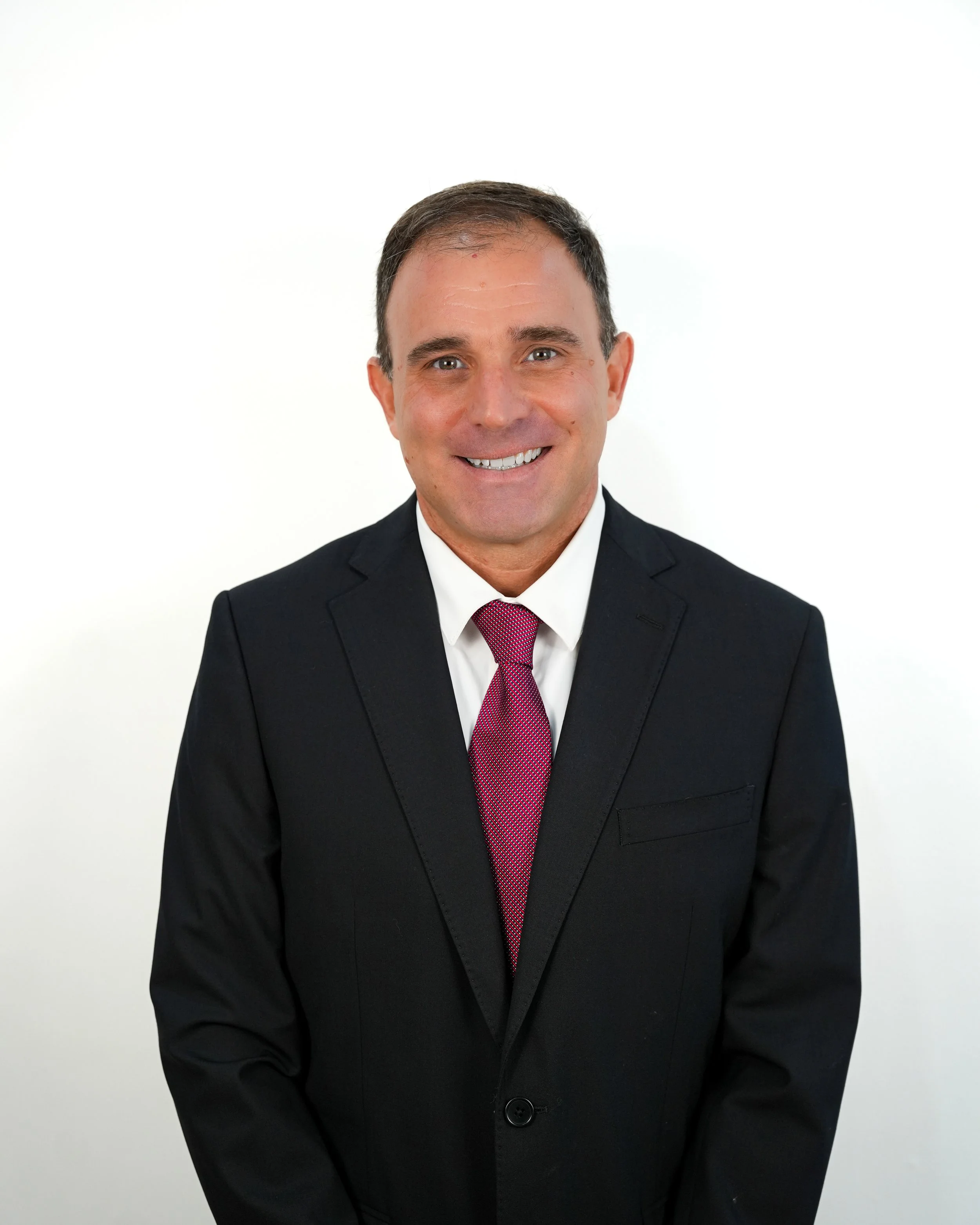 A man in a black suit, white shirt, and red tie, smiling, against a plain white background.