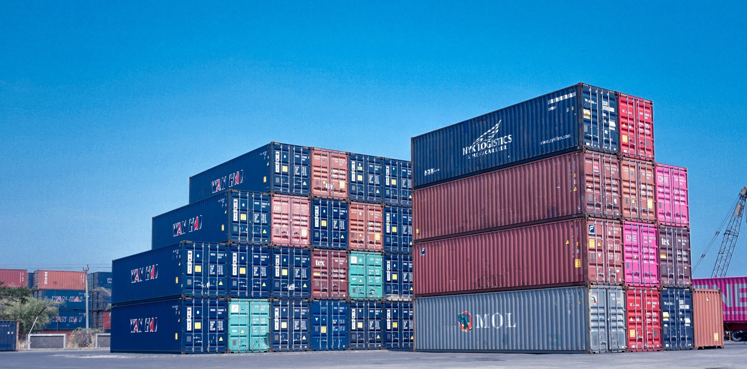 Multiple stacked shipping containers in blue, red, pink, and gray, with a crane in the background against a clear blue sky.