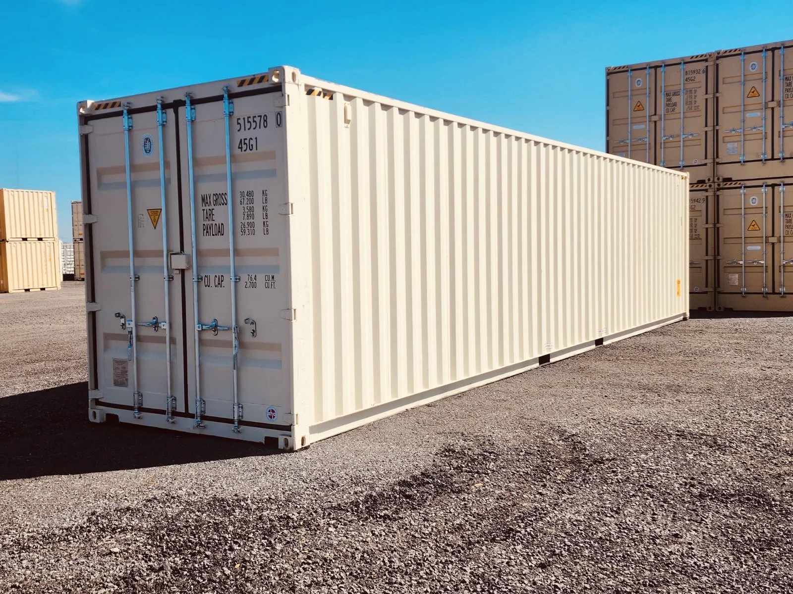 A large white shipping container standing on gravel with yellow and orange markings, set against a bright blue sky with other containers in the background.