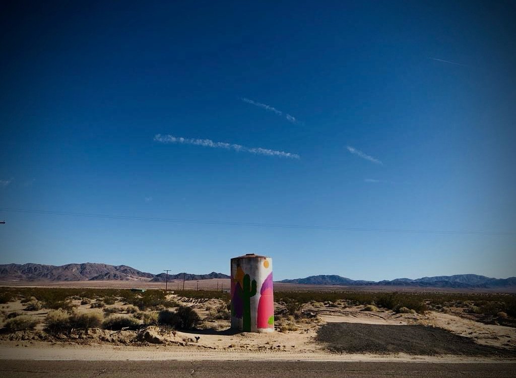 Photo of a water tower in Wonder Valley CA painted with a cactus and vibrant colors under a blue desert sky.