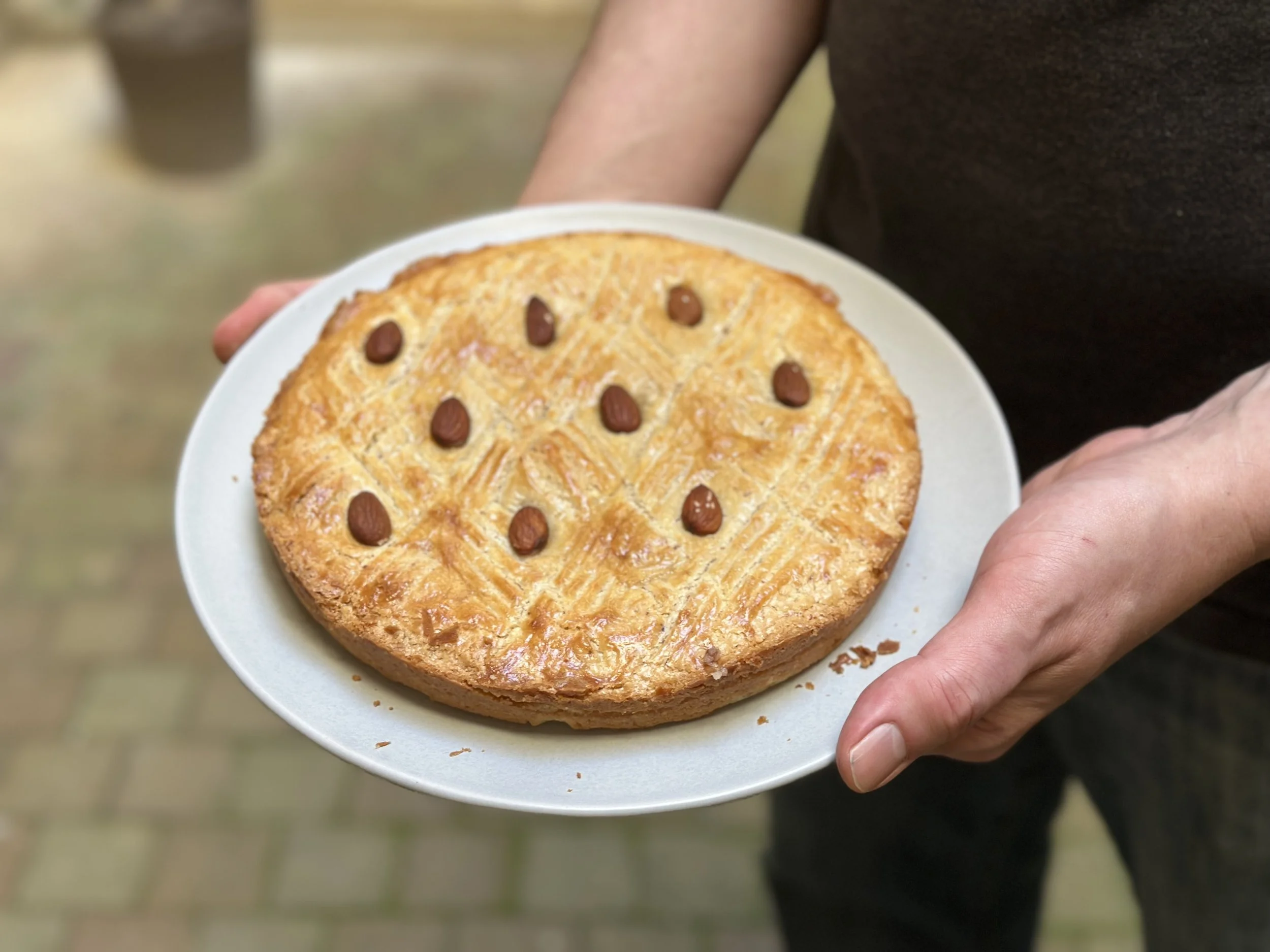 A person holding a white plate with a round, golden-brown pastry topped with almond slices and whole almonds. The background is blurred.