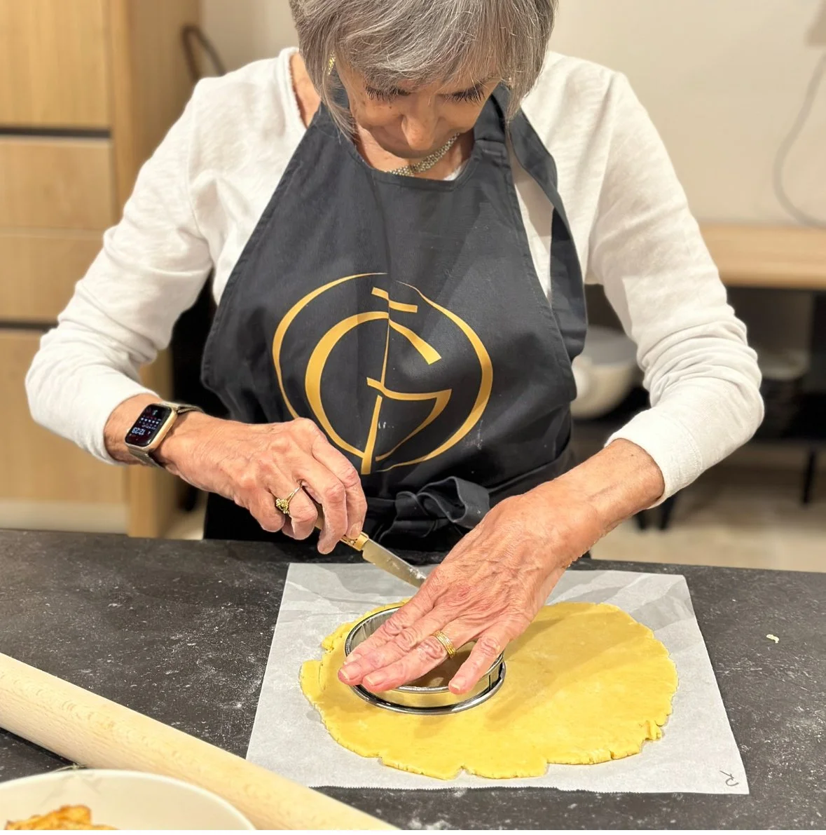 Older woman wearing a black apron with a gold logo, using a cookie cutter to cut dough, standing at a kitchen counter.