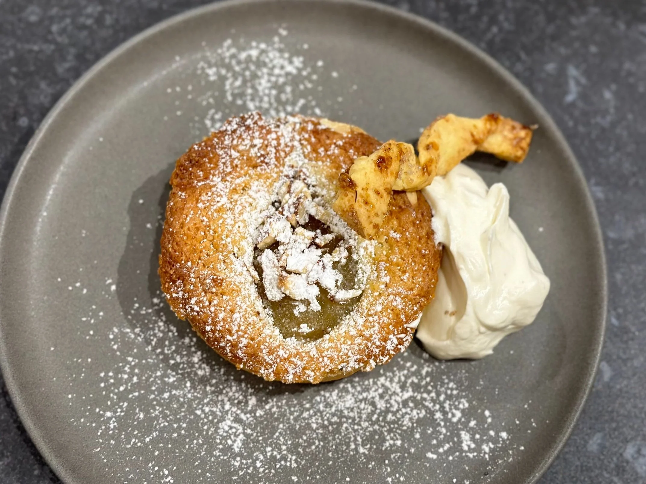 Apple dessert topped with powdered sugar, served with a side of whipped cream and a caramelized apple slice on a gray plate.