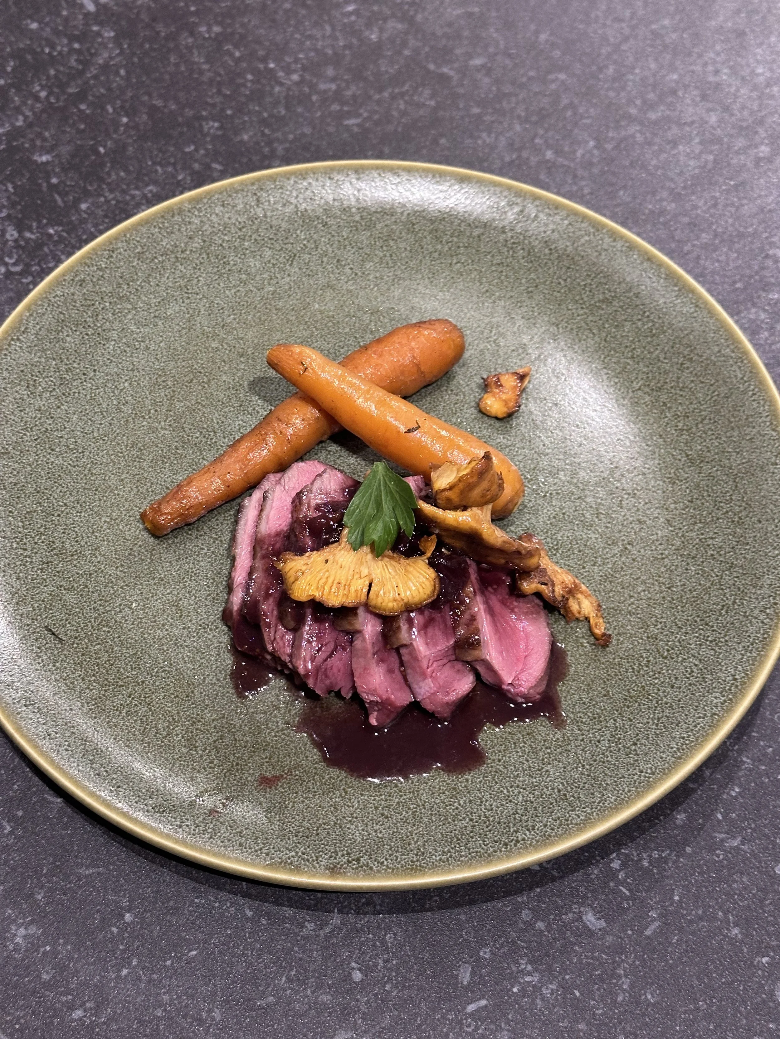 Seared duck breast with red wine sauce, roasted carrots, sautéed mushrooms, and a garnish of parsley on a green plate.