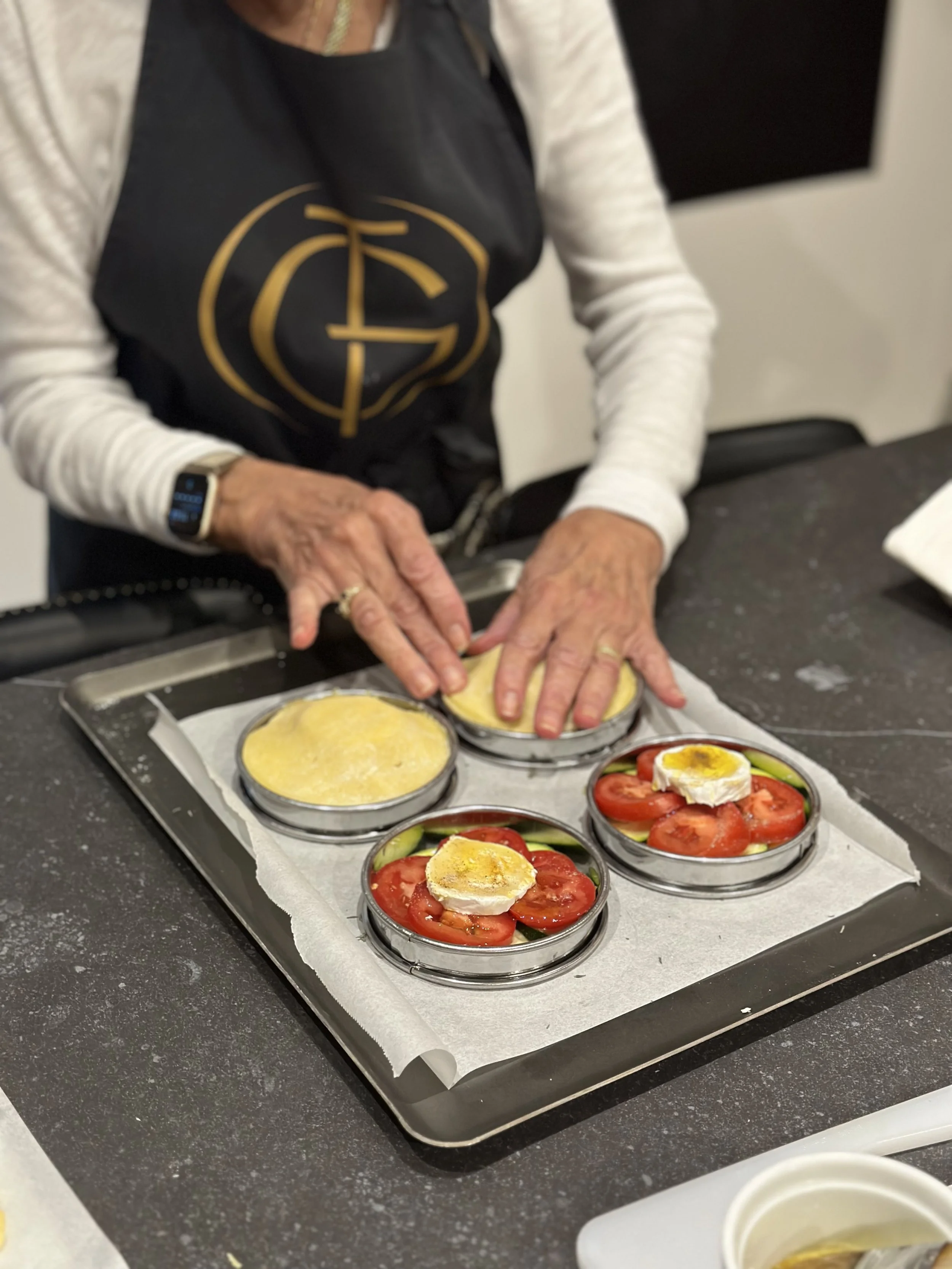 A person assembling individual tarts composed of tomato, zucchini, and goat cheese on a tray, with some layers covered with a yellow substance, in a home-styled teaching kitchen.