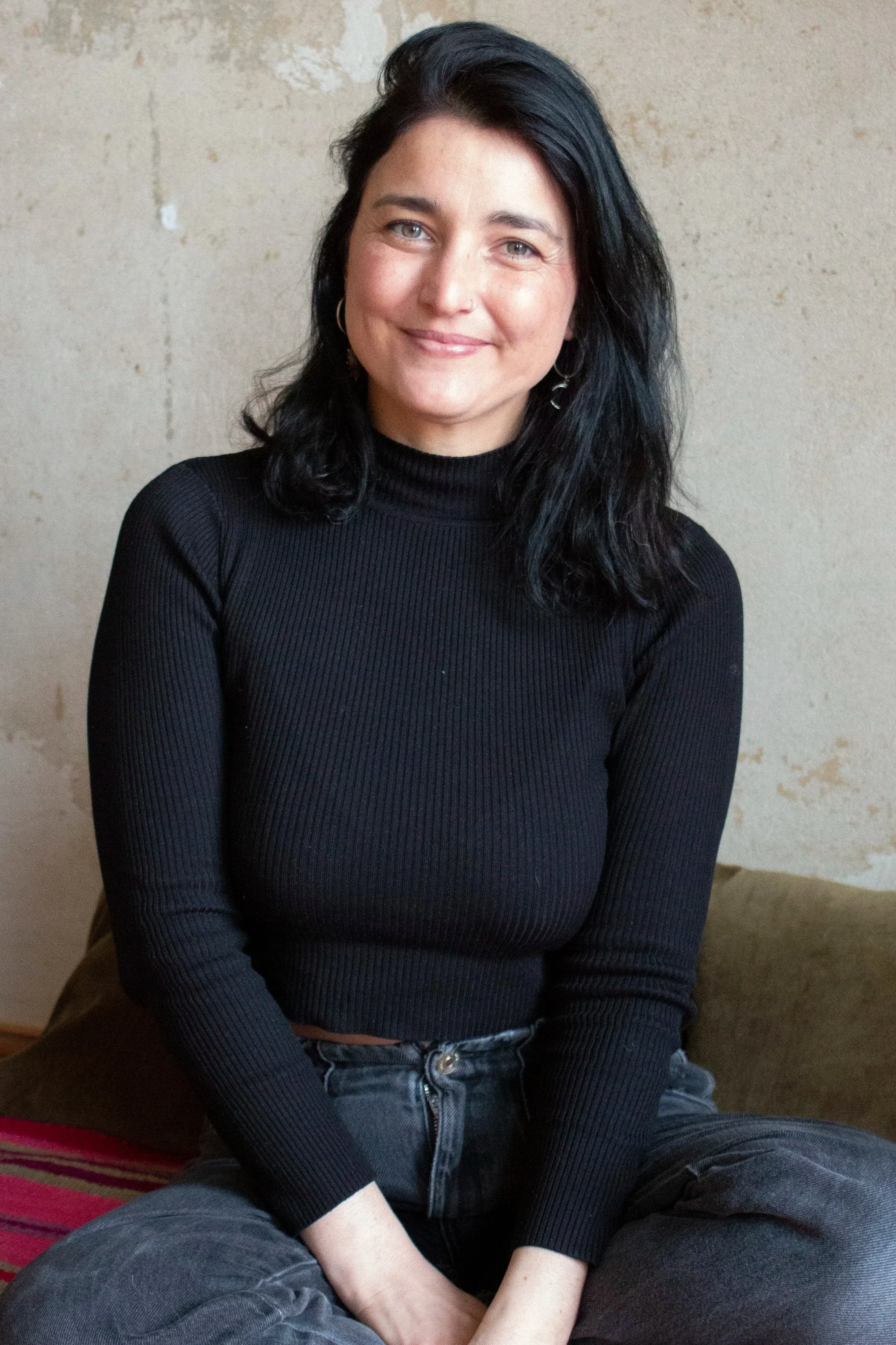 A woman with black hair and blue eyes, wearing a black ribbed turtleneck and gray jeans, sitting cross-legged against a textured beige wall and smiling at the camera.