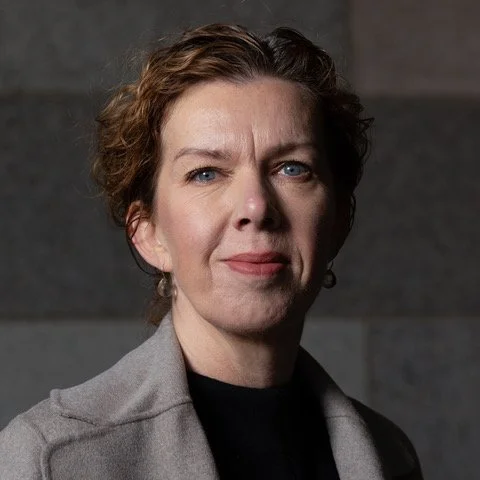 Portrait of a woman with short curly hair, wearing a gray blazer, standing against a gray wall.