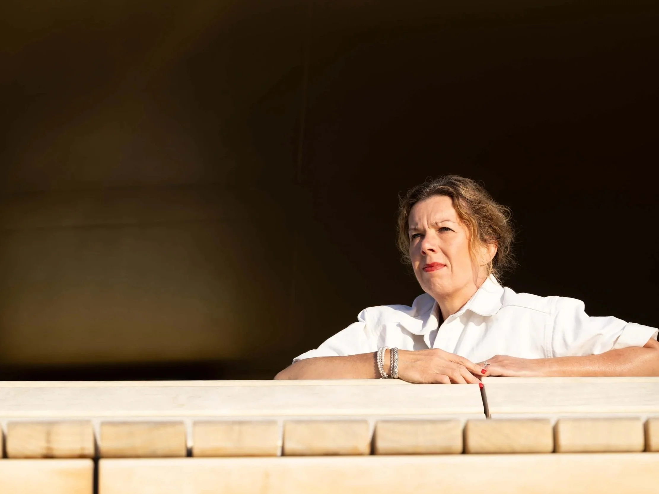 A woman with curly hair, wearing a white shirt, glasses, and jewelry, sitting outdoors with her arms resting on a ledge, looking thoughtfully into the distance against a dark sky.
