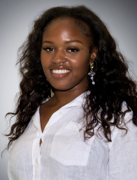 A woman with long, curly hair wearing a white shirt and blue earrings, smiling at the camera in front of a neutral background.