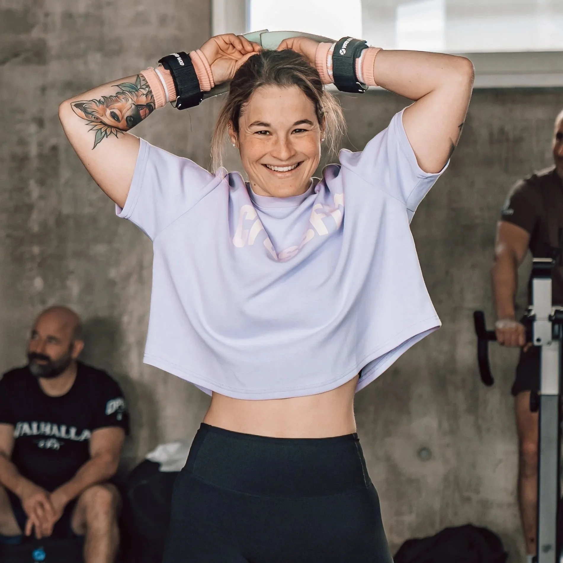 Une femme souriante en tenue de sport, tenant ses cheveux en arrière, avec un tatouage de renard sur le bras, dans une salle d'entraînement.