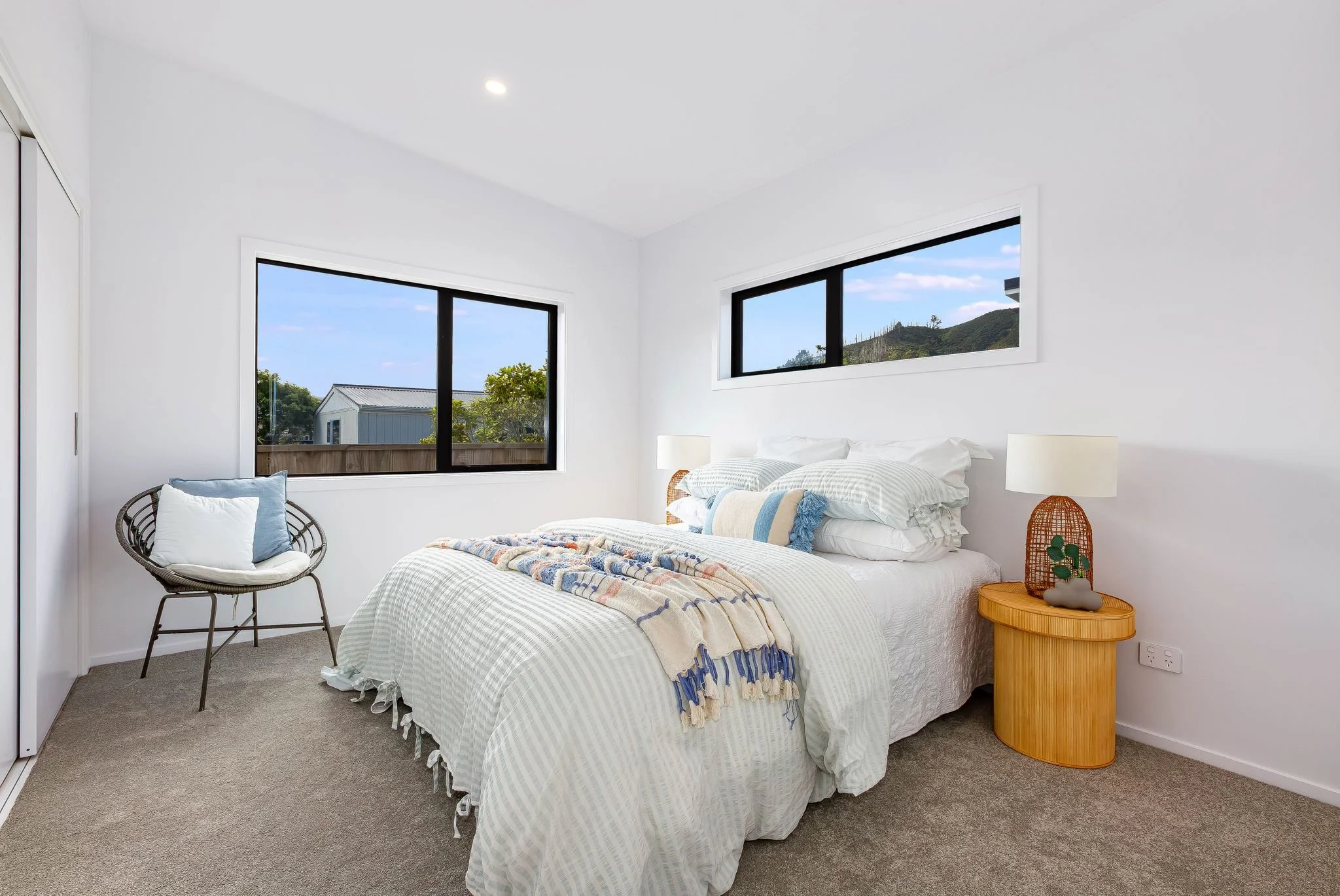 Bright bedroom with white walls, a bed with white bedding and blue accents, a round wooden bedside table with a lamp, a wicker chair with cushions, and large windows showing a blue sky and outdoor trees.