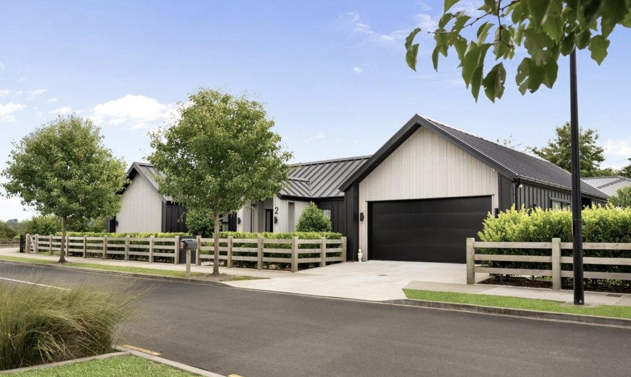 O'Brien Homes: A modern residential house with a black and white exterior, a pitched metal roof, and a two-car garage, surrounded by trees and a wooden fence along a paved street in a suburban neighborhood.