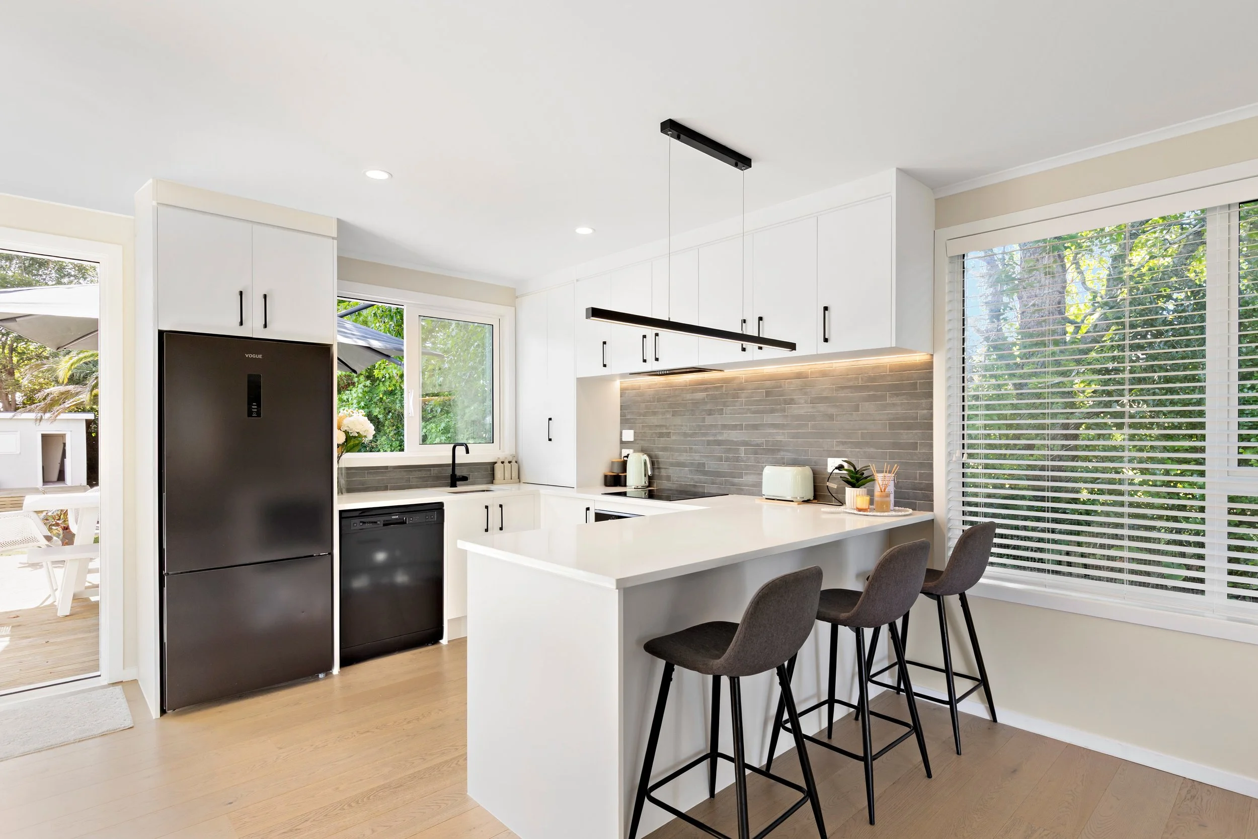 Modern kitchen with black refrigerator, white cabinets, gray backsplash, and large window with blinds overlooking greenery.