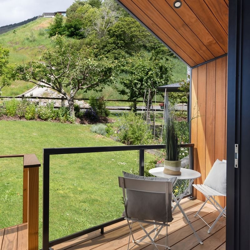 View of a balcony overlooking a garden with trees and greenery. The balcony has a small white table with a potted plant, and two white folding chairs. The ceiling and wall of the balcony are wood-paneled, and there is a glass railing.