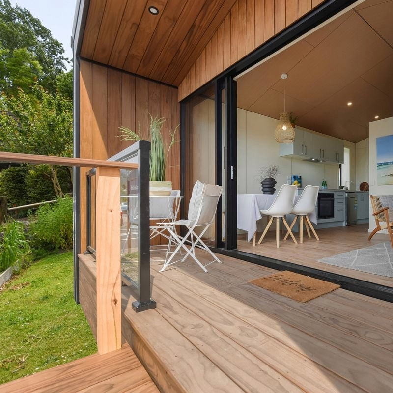 View from a wooden balcony of a modern house, showing an open sliding door, interior kitchen and dining area, and lush green outdoor space.