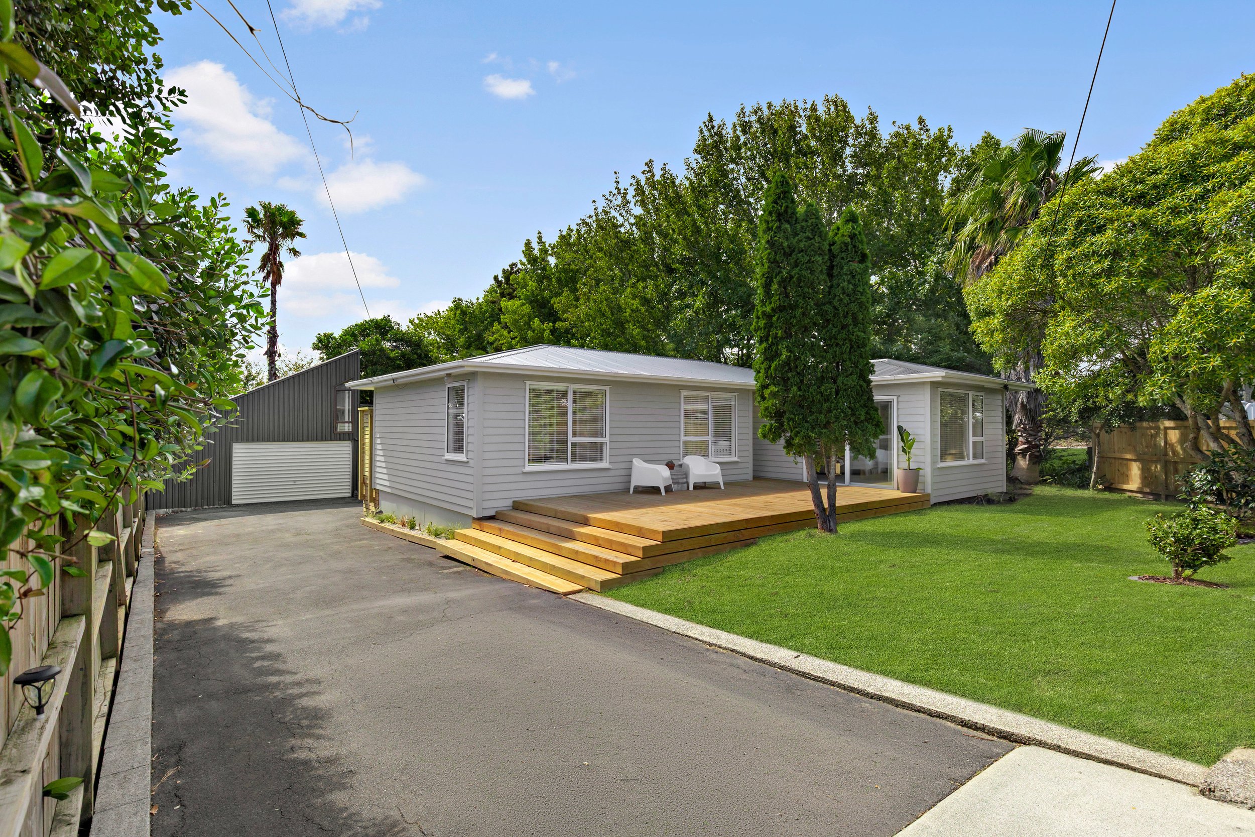 A white house with a wooden deck and stairs, green lawn, trees, and a driveway leading to a metal garage, under a partly cloudy sky.