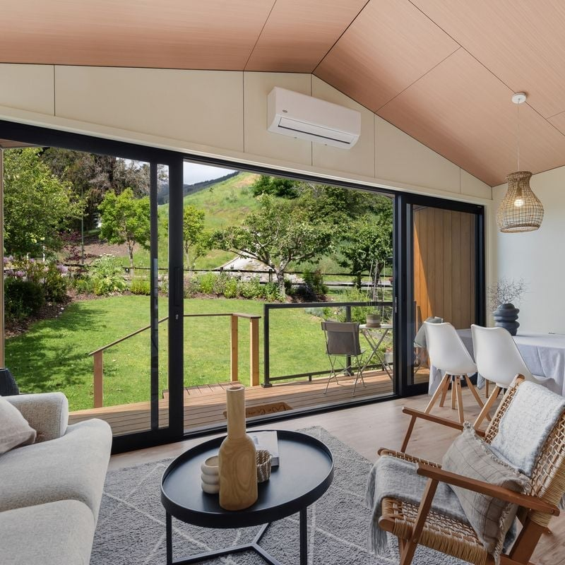 Living room with large glass sliding doors overlooking a green yard and hill, furnished with a beige sofa, wicker chair with cushion, white dining chairs, and a black round coffee table with vases and small decorative items, patterned rug, and wooden