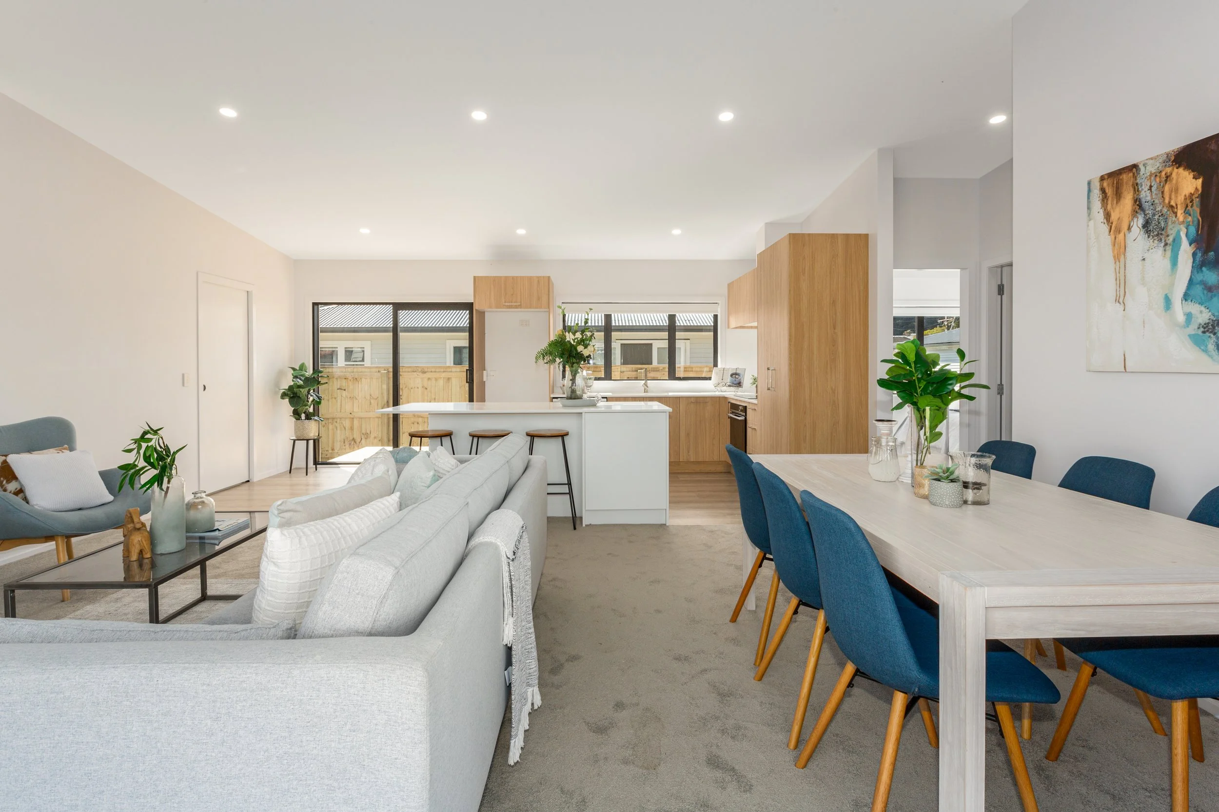 Open-concept living and dining area with a gray sofa, wooden dining table with blue chairs, and a modern kitchen with white countertops, wooden cabinets, and large windows letting in natural light.