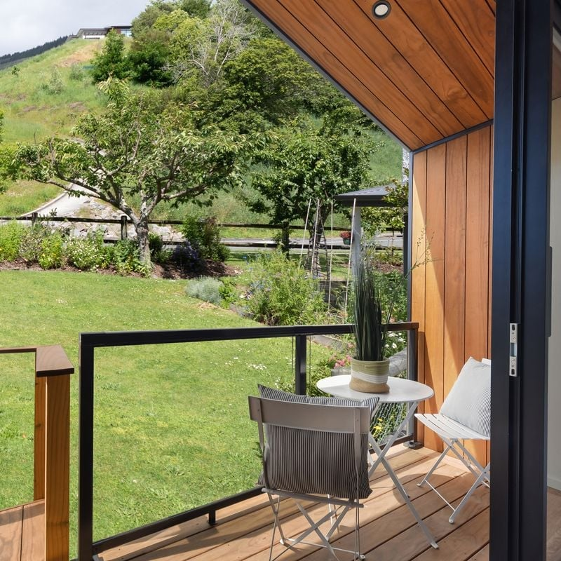 View of a small balcony with two metal chairs, one with a cushion, and a round table with a potted plant, overlooking a landscaped yard with trees and a grassy hill in the background.