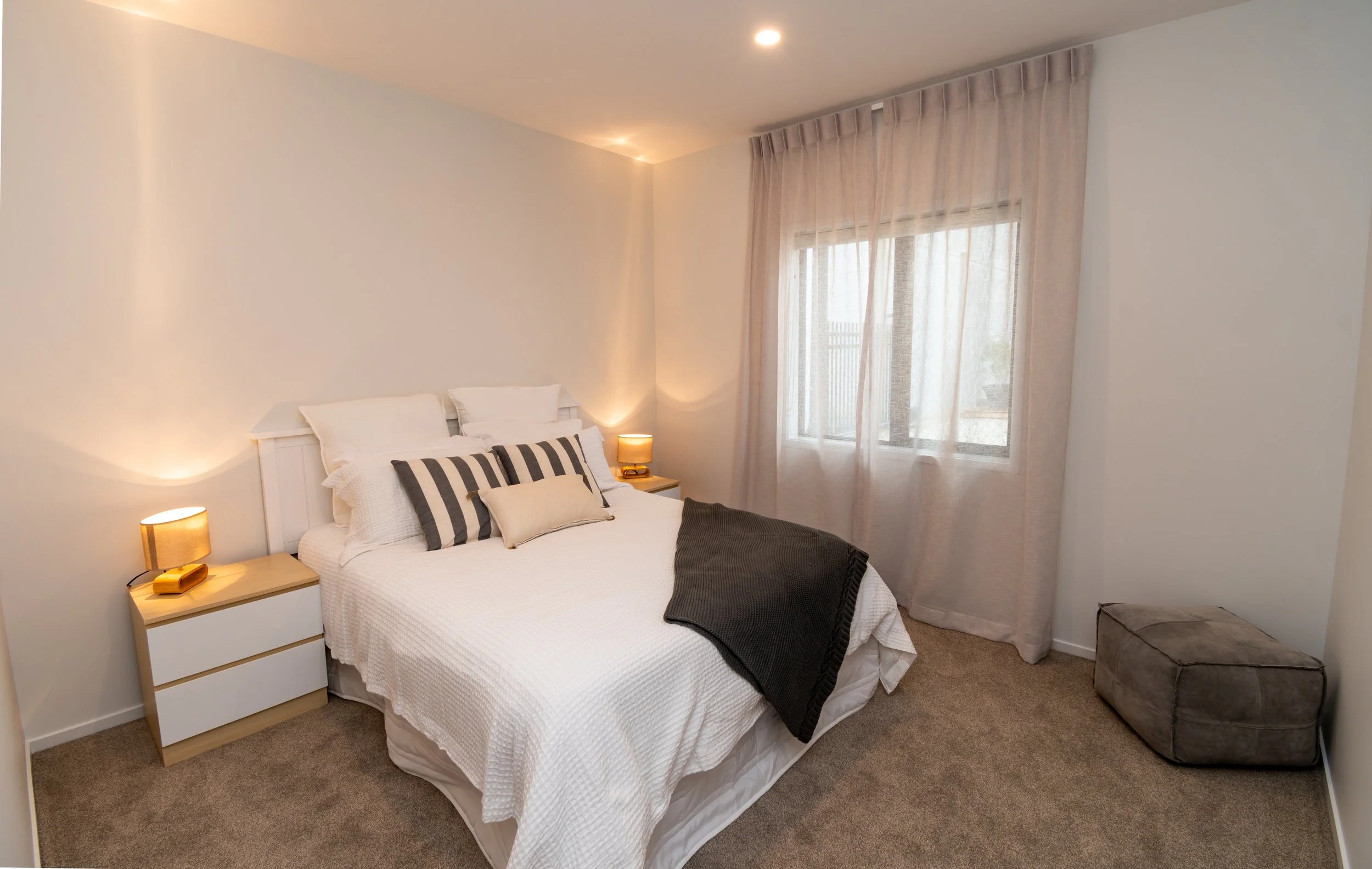 A neatly made bed with white bedding and decorative pillows in a bedroom with bedside lamps, a window with light curtains, and a brown ottoman.