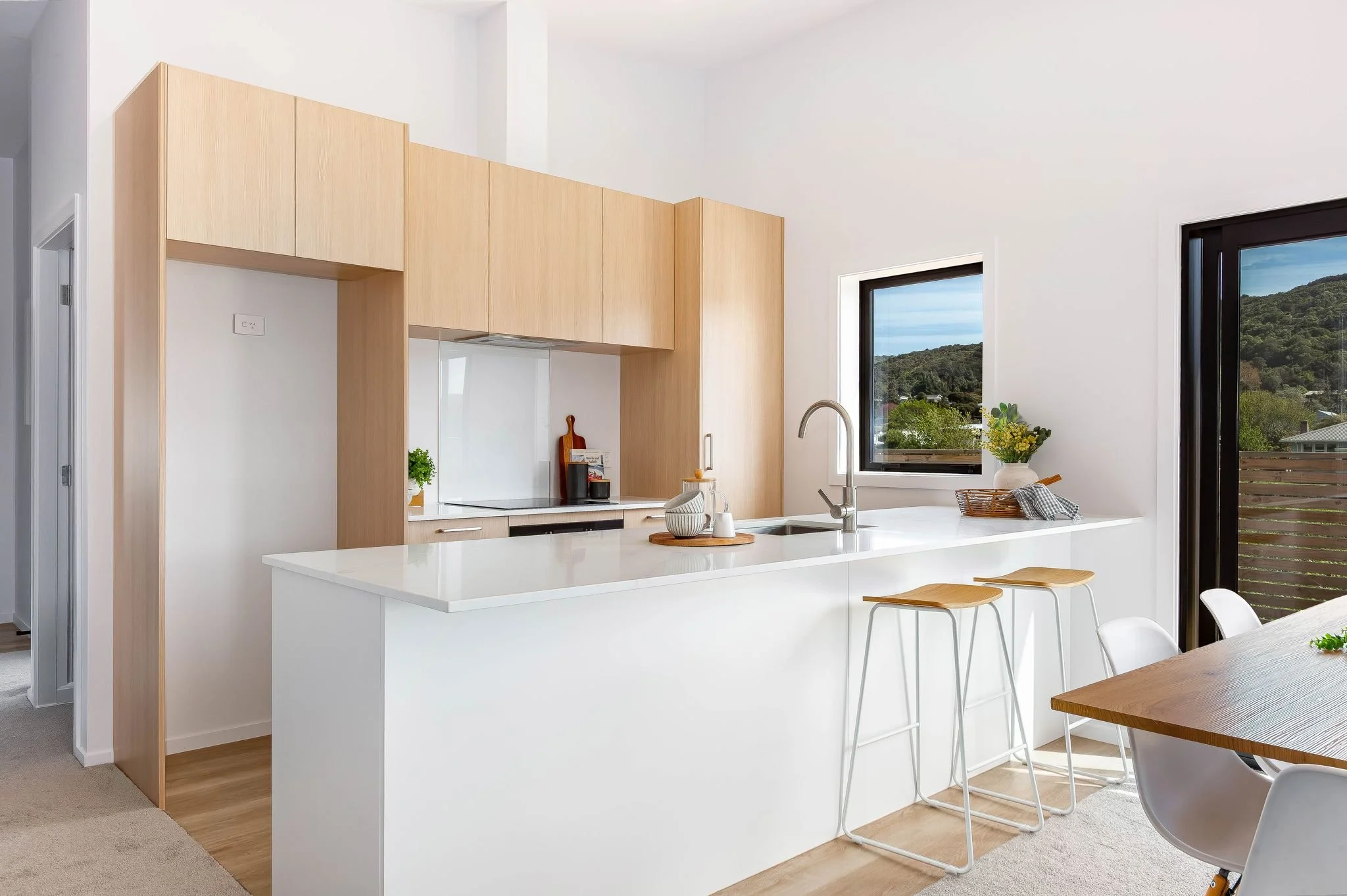 Modern kitchen with white island, wooden cabinets, black framed windows overlooking greenery, and a wooden dining table with white chairs.