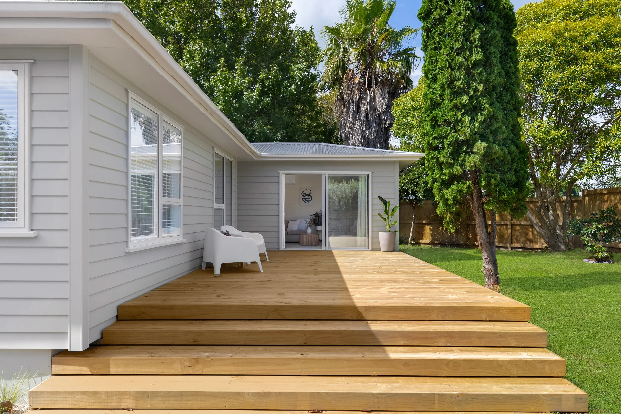Backyard with wooden deck, white house with multiple windows, sliding glass door leading to interior, outdoor seating area with two white chairs, potted plant, tall trees, and lush green lawn.