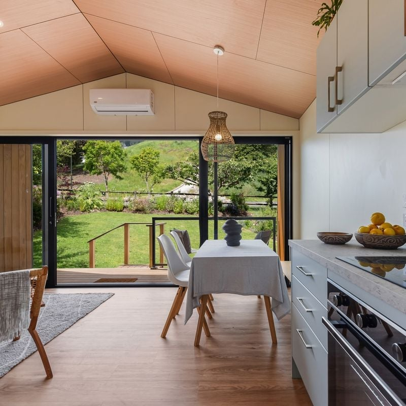 Interior view of a bright dining area with a view of a green garden through large sliding glass doors, featuring a wooden ceiling, white walls, modern furniture, and minimal decor.