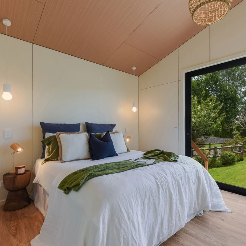 Cozy bedroom with a bed, multiple pillows, green and white blankets, and hanging pendant lights, with a large sliding glass door opening to a green yard.