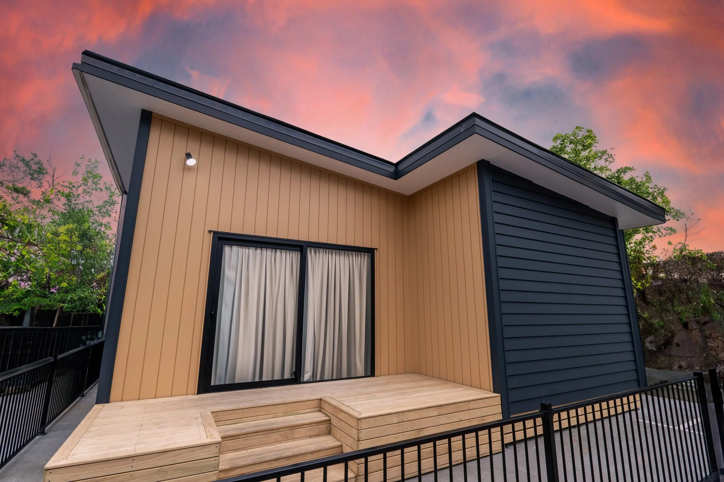 Clever Living Co. A modern house with blue and beige siding, a sliding glass door with curtains, and a small wooden porch at sunset with a colorful sky and trees in the background.