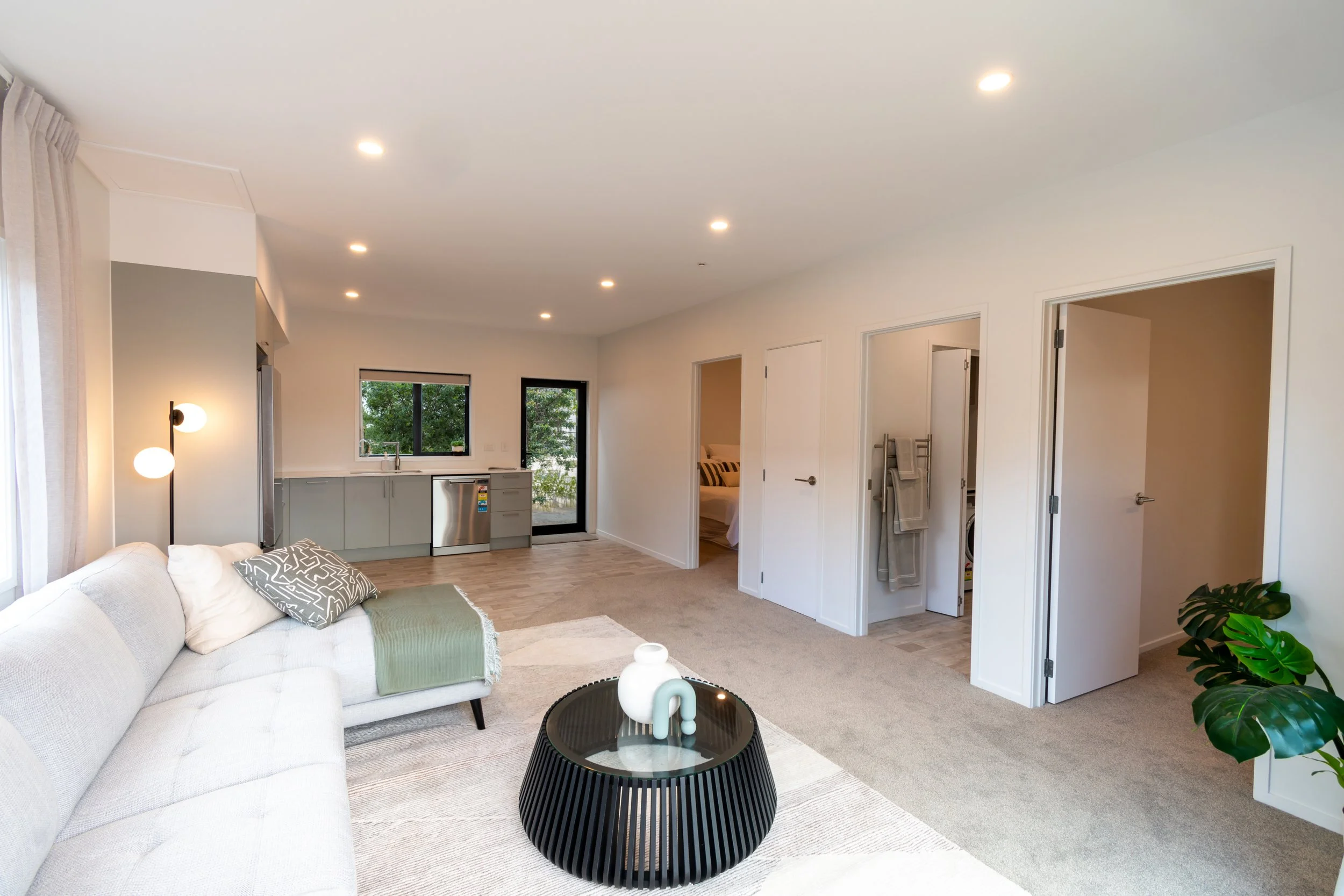 Living room with white couch, black coffee table with decorative items, kitchen area with sink and cabinets, doors leading to two bedrooms and a laundry room, potted plant, modern decor, large windows, and recessed lighting.