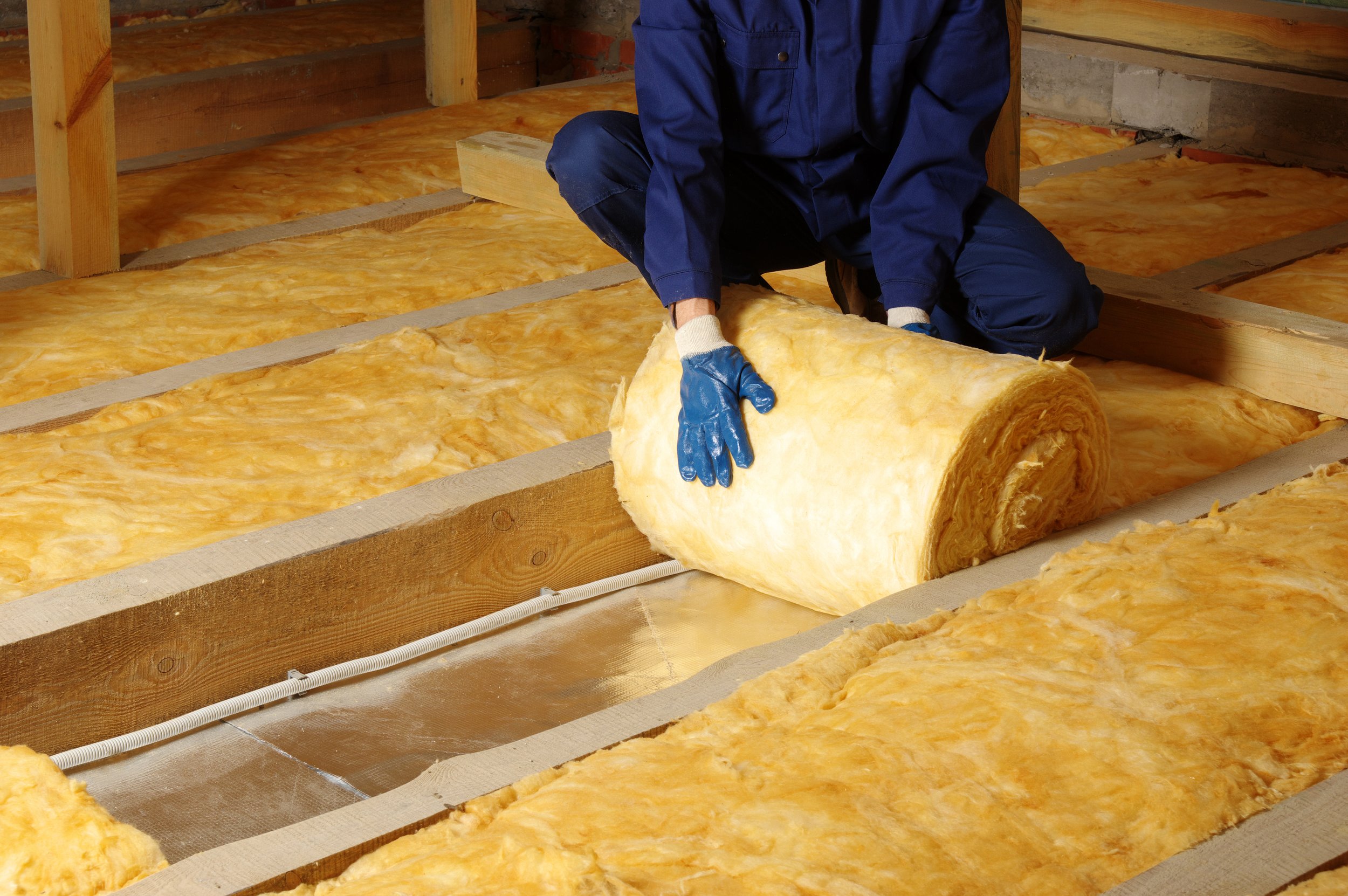 A person wearing blue overalls and gloves is rolling out and installing loft insulation in an attic or loft. In the UK.