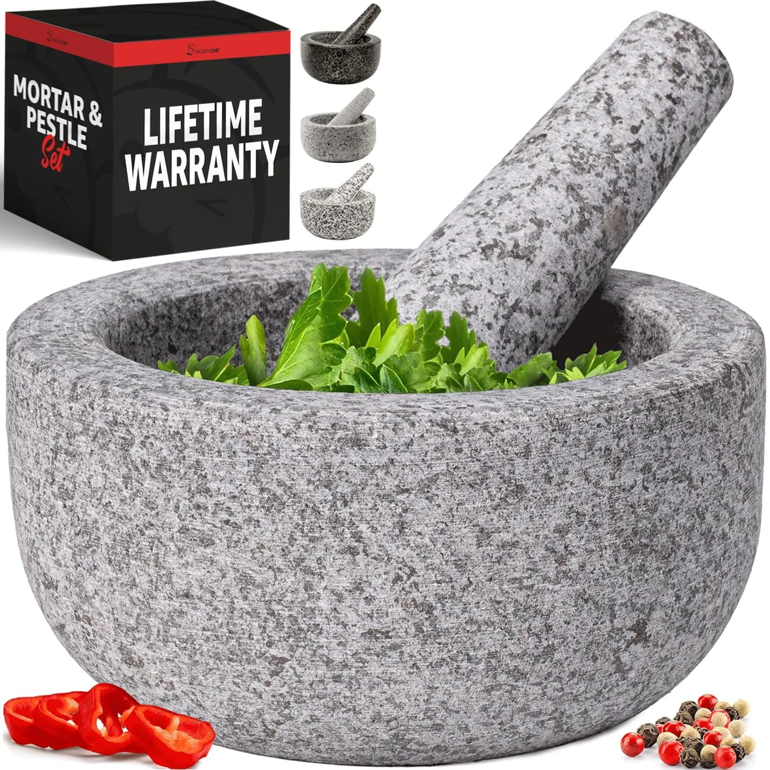 A close-up of a gray granite mortar and pestle with fresh green herbs inside. In the background, there are three smaller mortar and pestle sets and a black box labeled 'Mortar & Pestle Set' with a lifetime warranty. In the foreground, there are slices of red chili pepper and a mix of peppercorns.