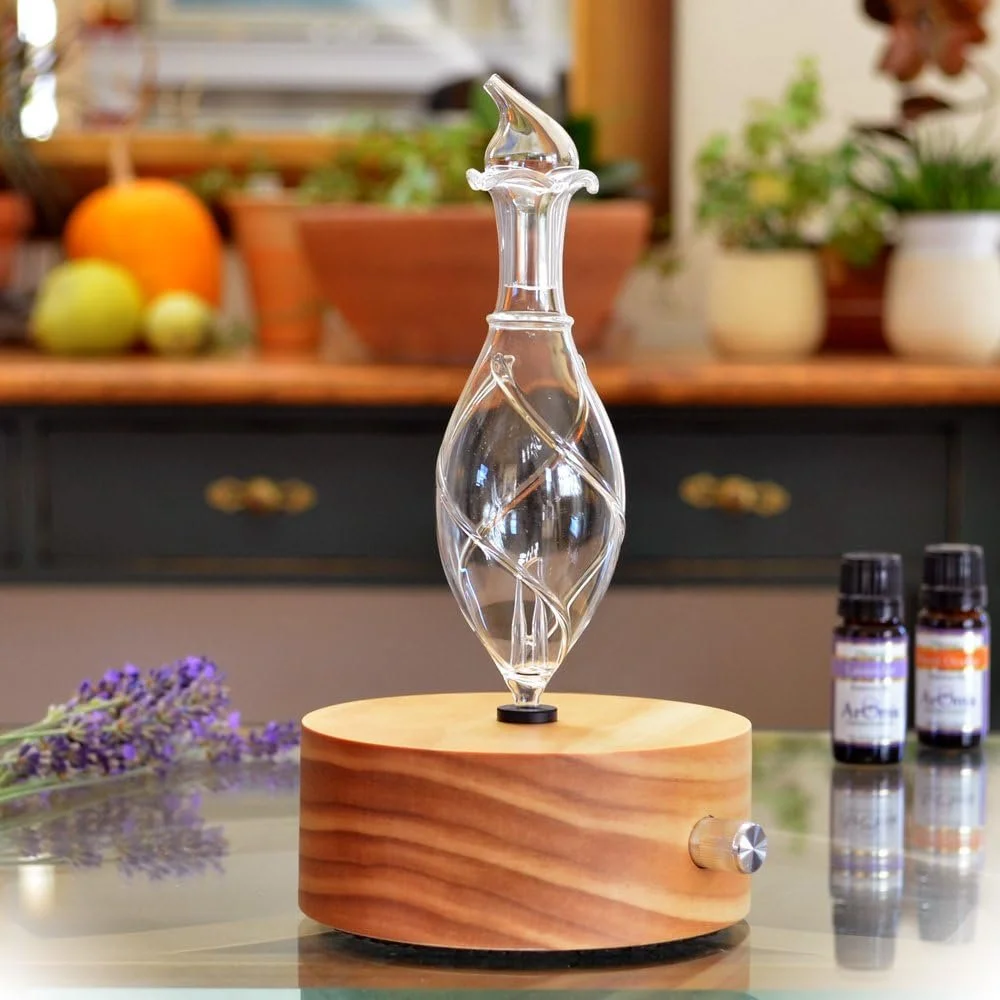 A glass diffuser on a wooden base, with two essential oil bottles and lavender sprig on a glass table in a kitchen.