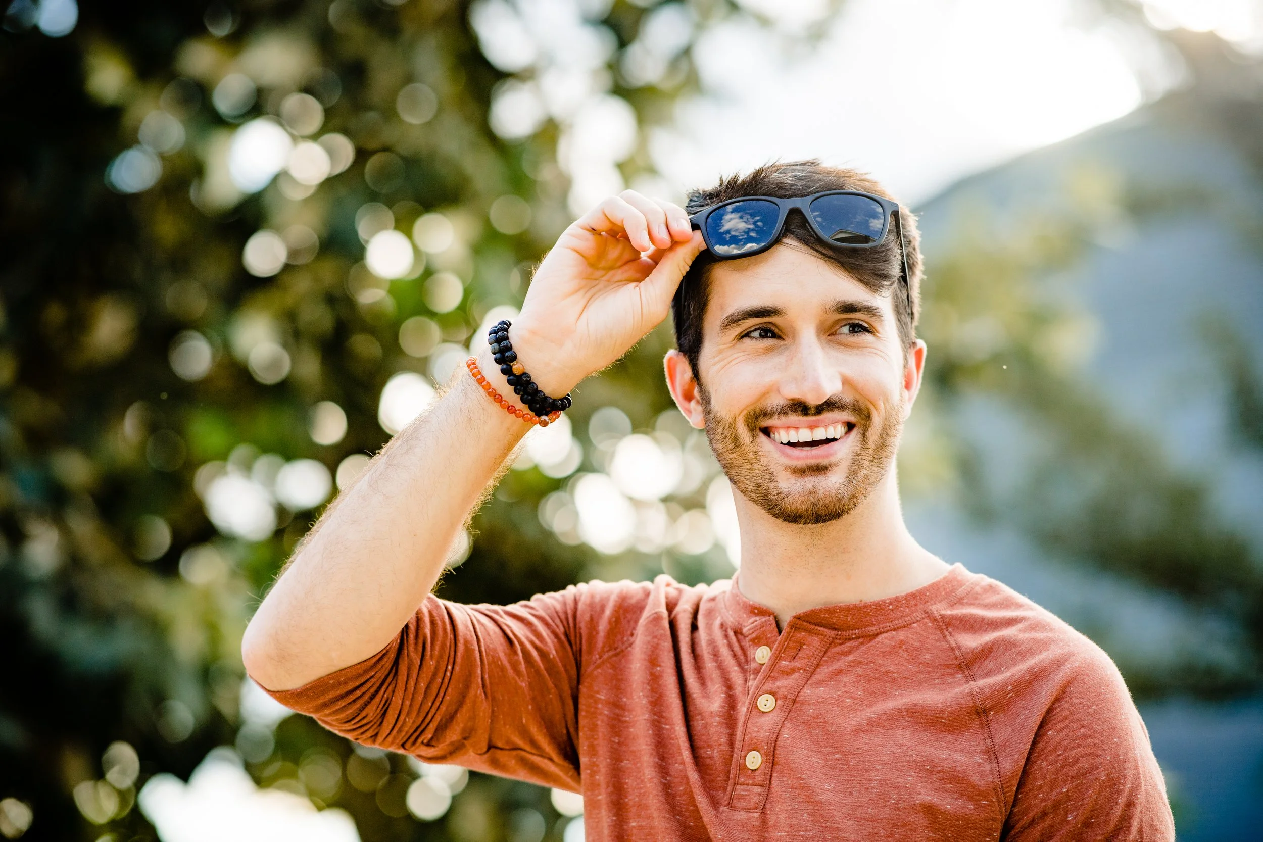 Man smiling outdoors, holding sunglasses on his head, wearing a reddish-brown shirt and beaded bracelets.