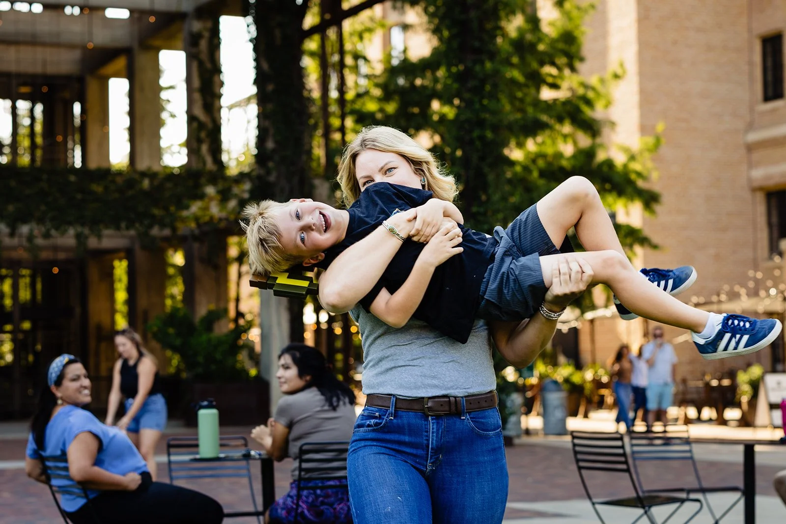 A woman lifting a young boy in her arms outdoors at a park or street scene, smiling, with other people seated and socializing in the background.