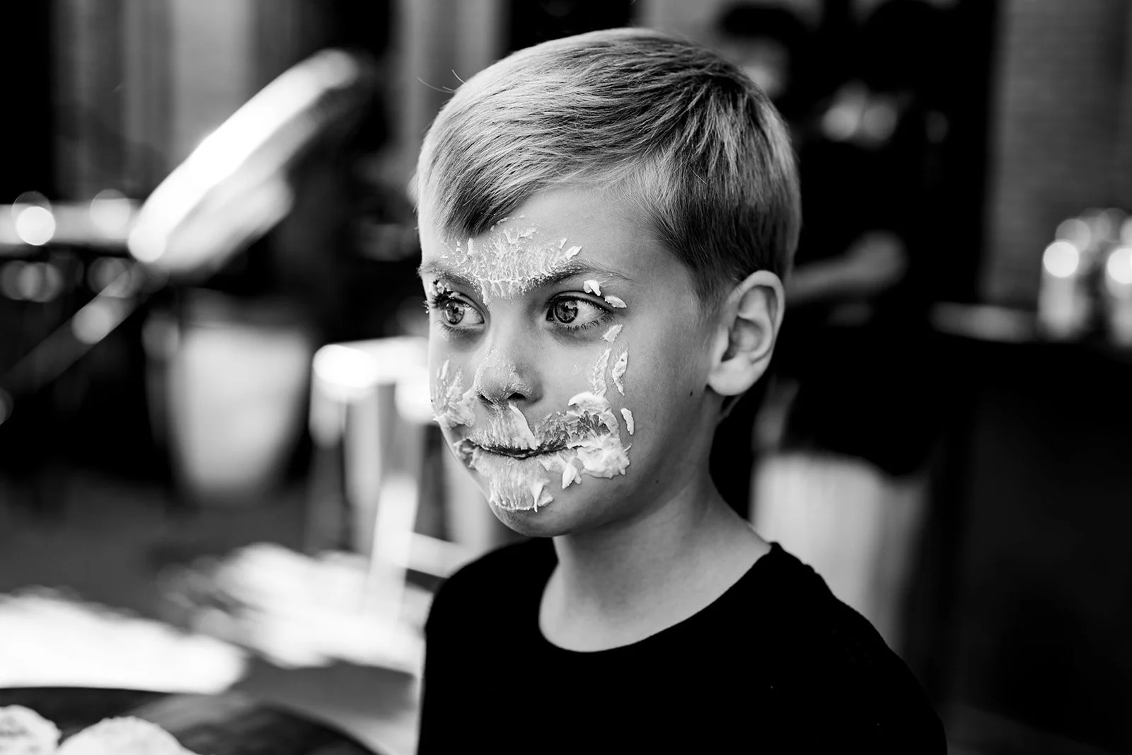 A boy with short hair has cake or frosting on his face, mostly on his forehead, nose, and mouth, in a black-and-white photo. There are blurred objects in the background.