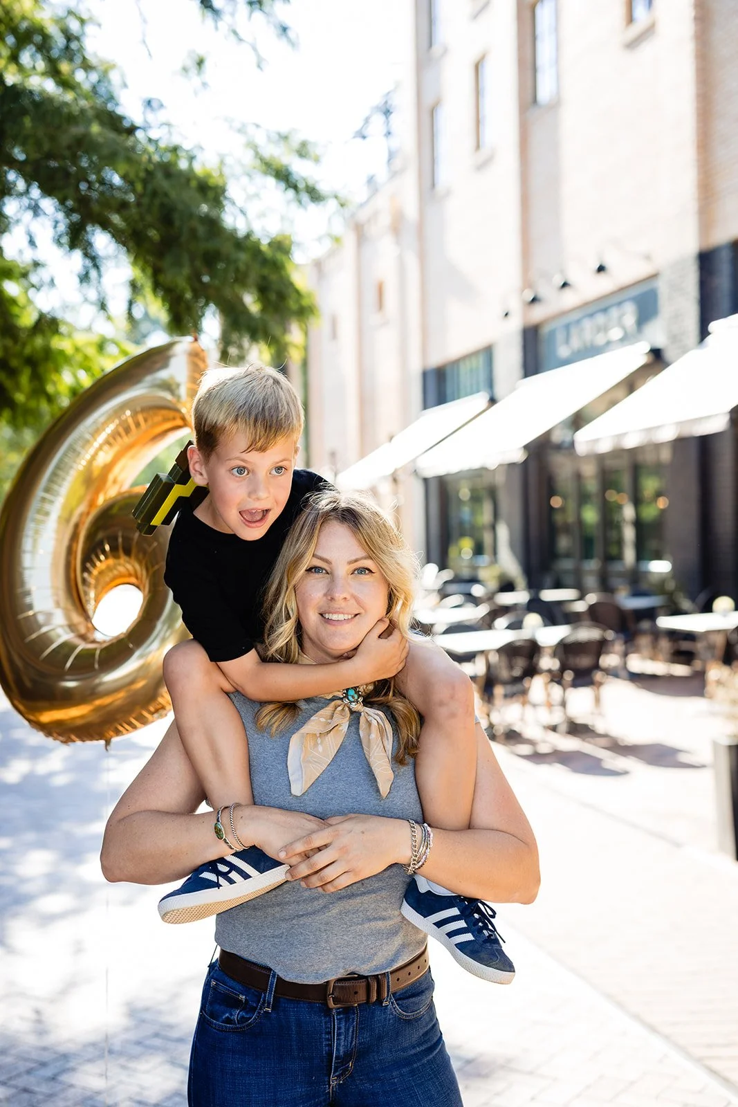 A woman giving a piggyback ride to a young boy outside on a sunny street with storefronts and outdoor seating, holding a large gold balloon shaped like the number 0.