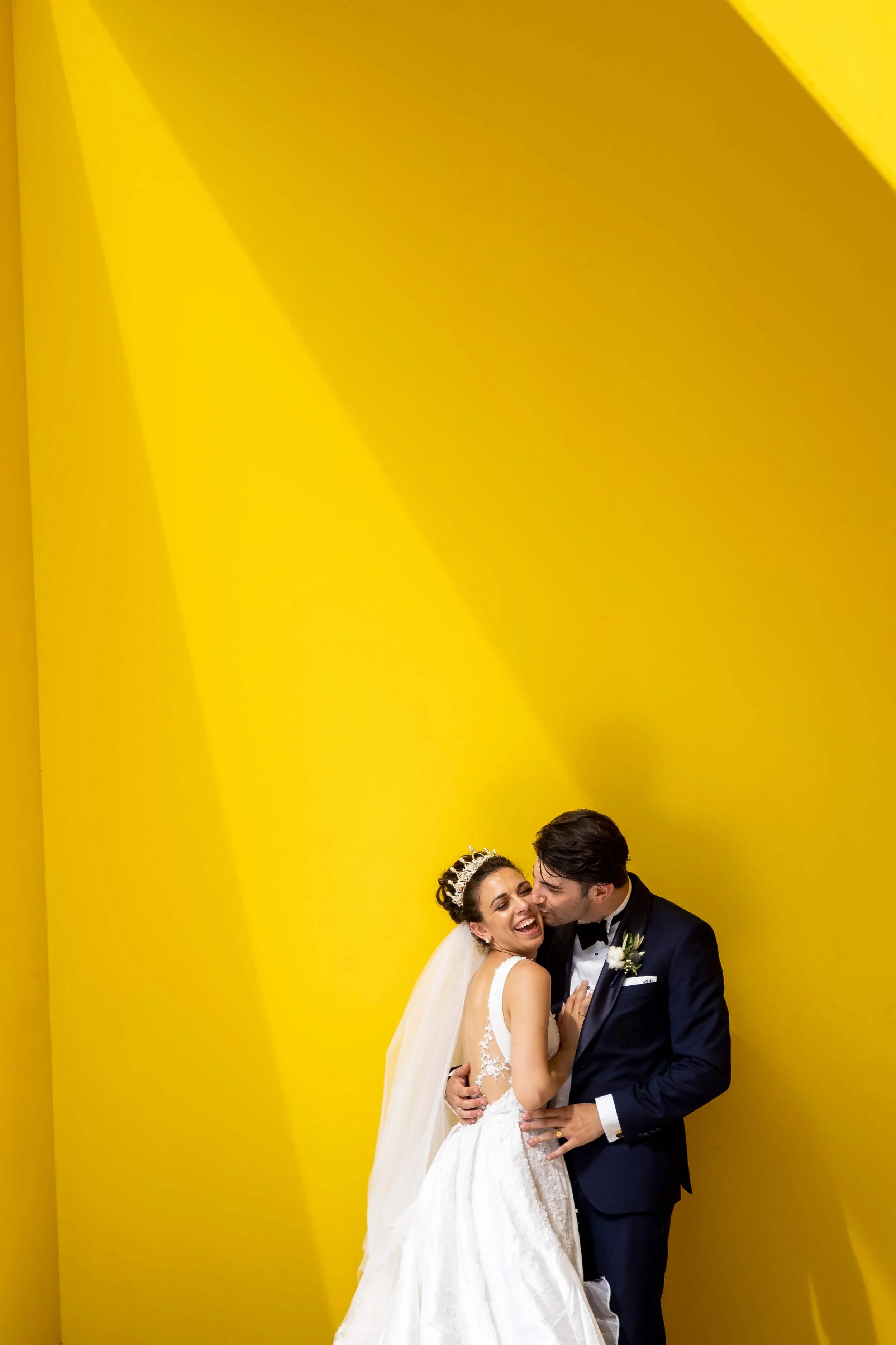 A bride and groom dancing and laughing together against a vibrant yellow background.
