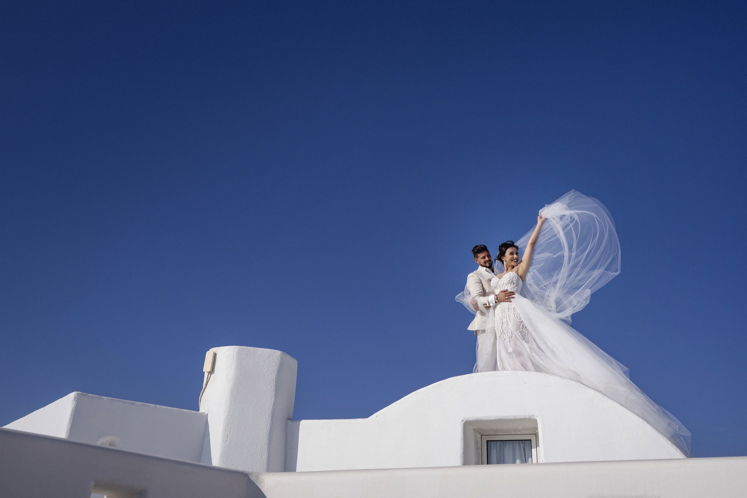 A wedding couple dressed in white on a rooftop with a clear blue sky background. The bride is holding her veil and smiling, while the groom stands beside her, holding her waist.