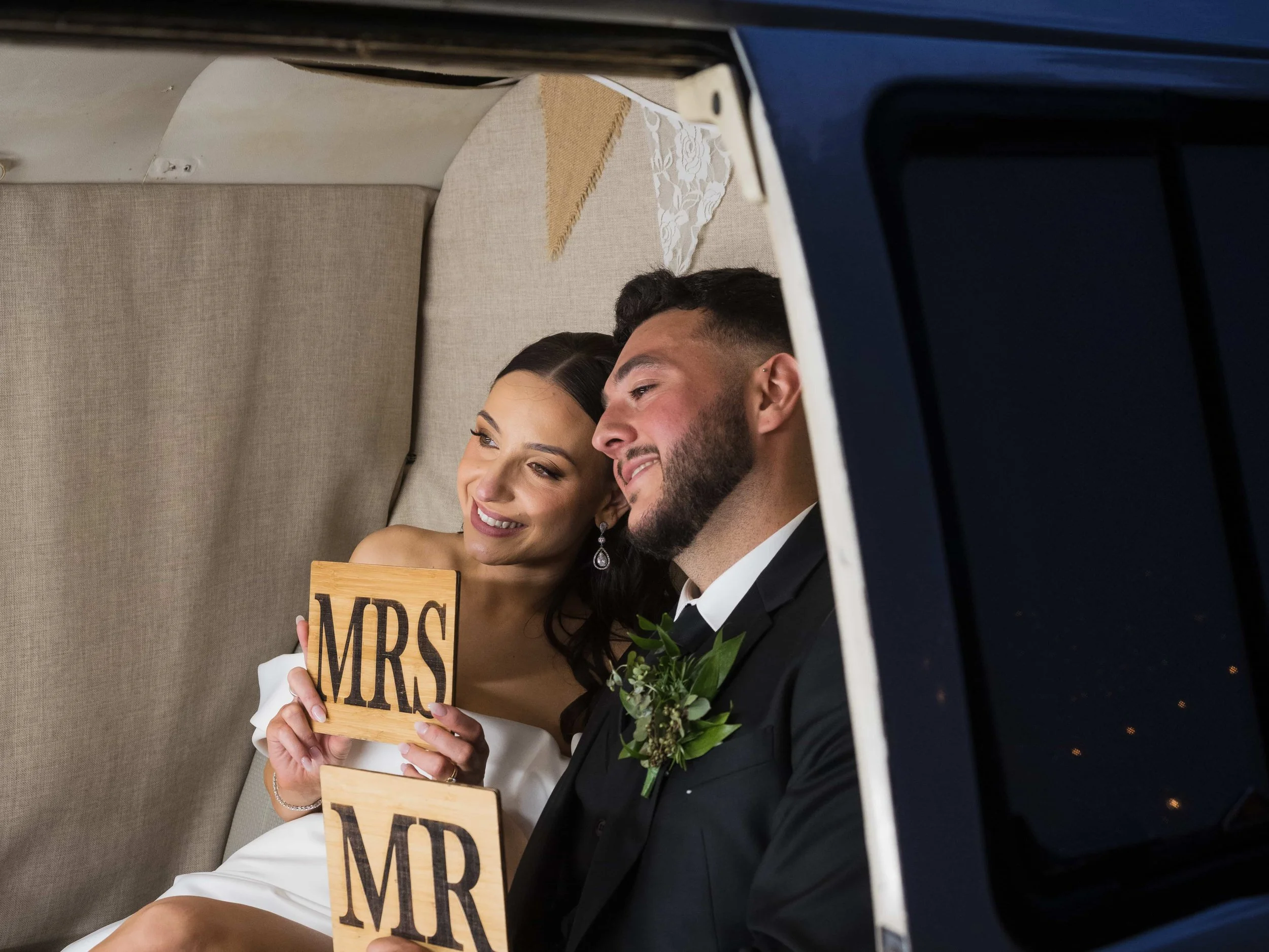 Happy bride and groom sitting together, holding wooden signs that read 'MRS' and 'MR', smiling and looking at each other, inside a vehicle.