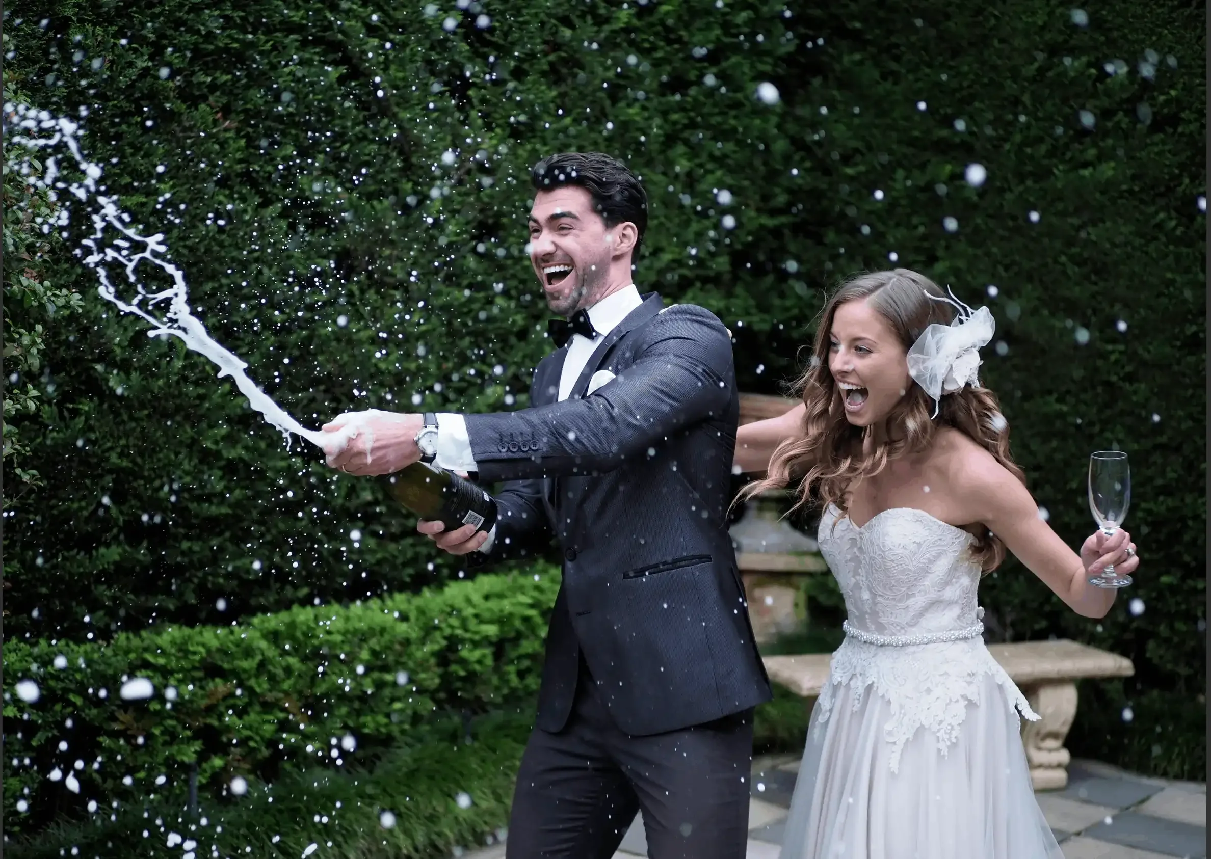 A newlywed couple celebrating outdoors with champagne. The groom is in a tuxedo and the bride in a wedding dress, both excited as champagne sprays around them.